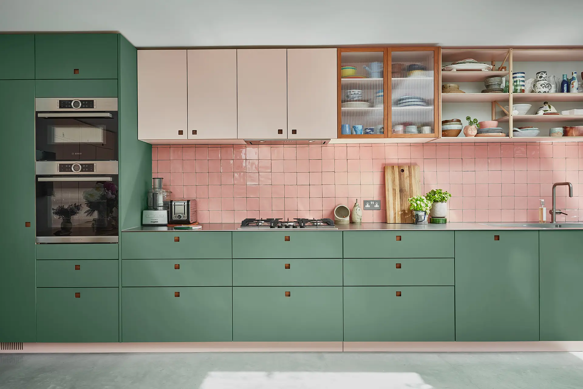 Pink and green Pluck kitchen cabinetry including a multi-coloured larder with sleek stainless steel worktops and glossy tiles.