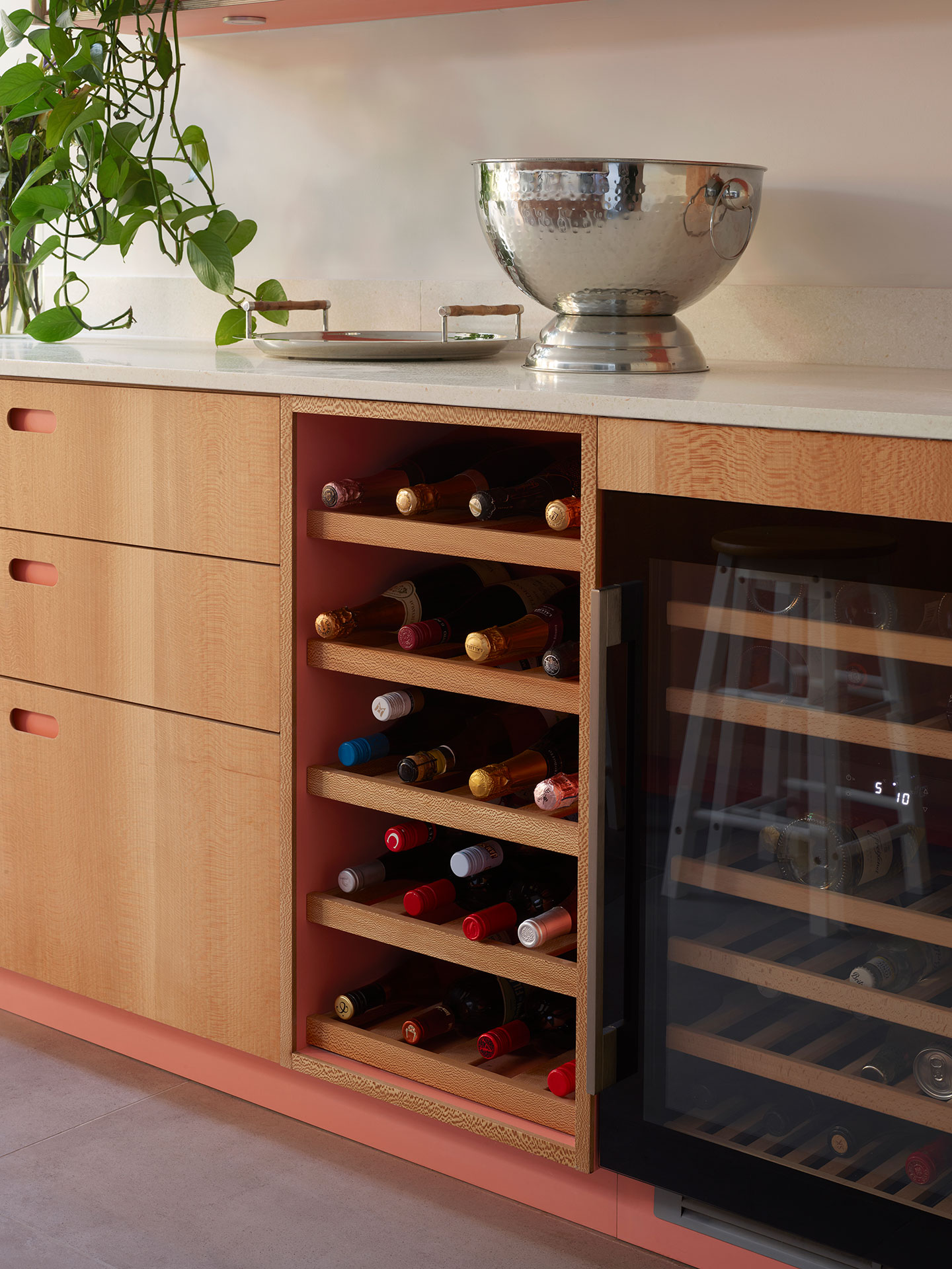 A wine rack in Dark grey and coral pink open plan kitchen with a London Plane wood cocktail cabinet and waterfall edge terrazzo topped island.