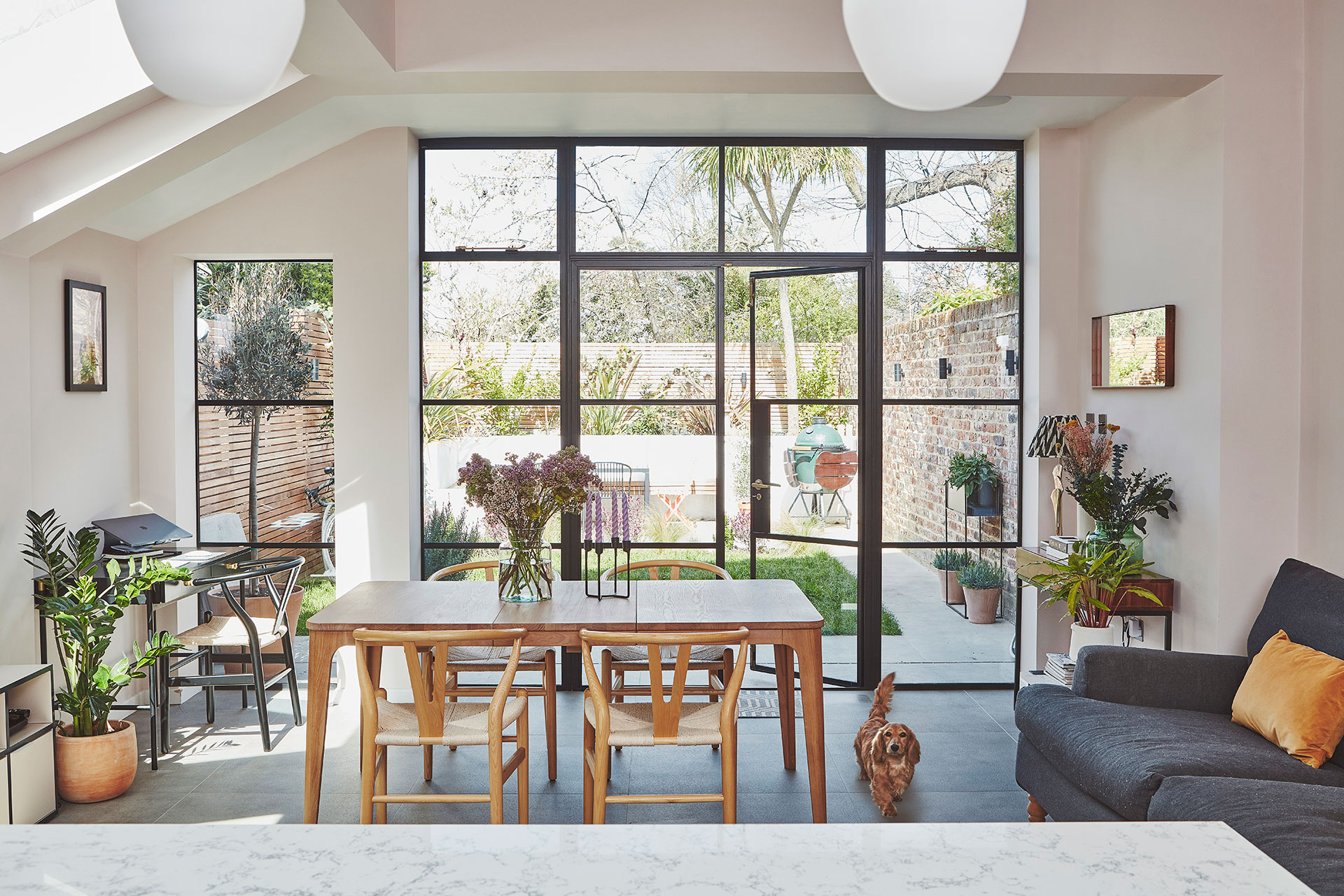 Pluck's white and wood kitchen cabinetry topped with mottled natural stone quartz, a dining table, chairs and sofa.