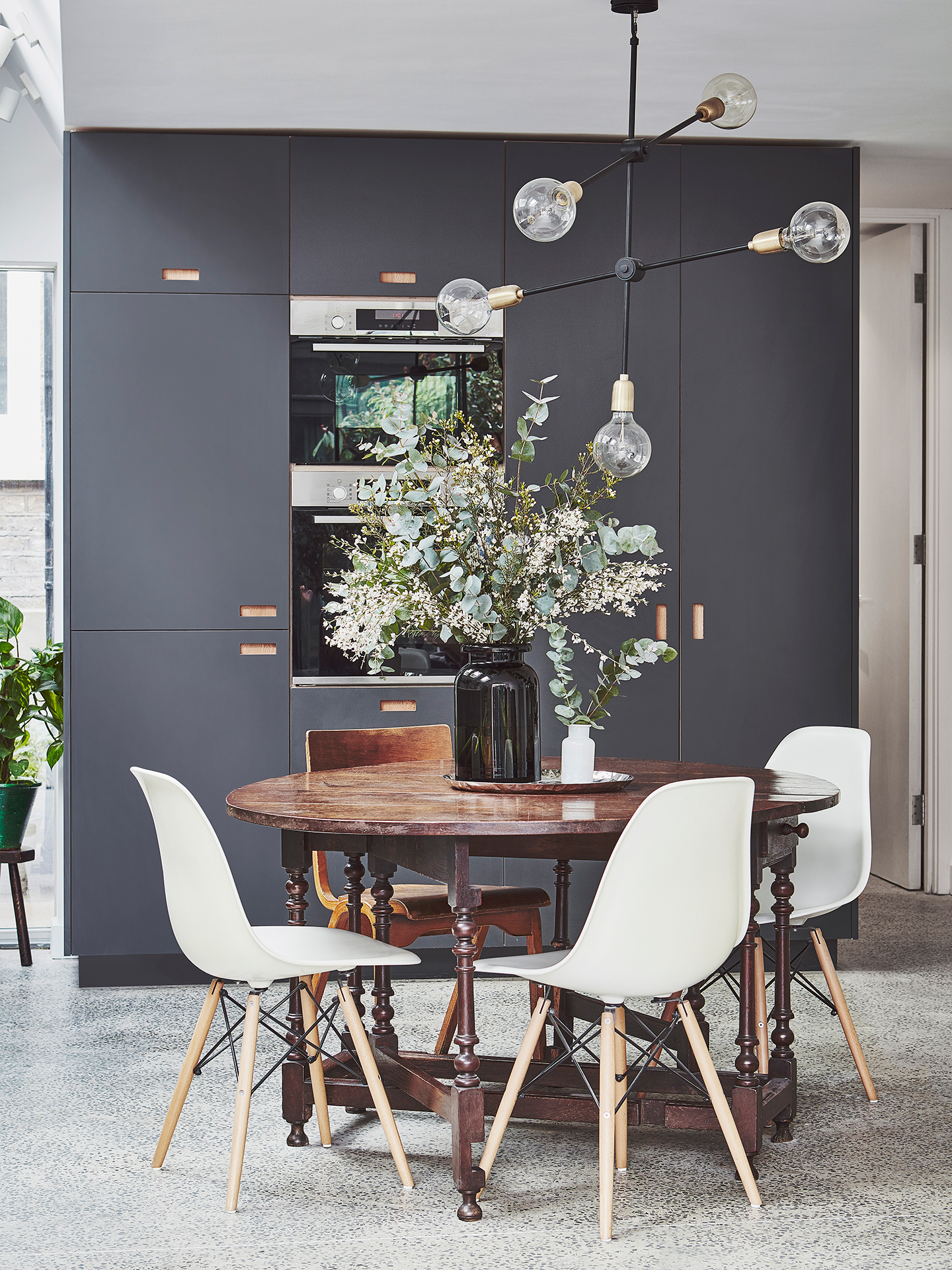 A glass pendant sits above a round dining table with white chairs. Behind, there are tall grey kitchen cabinets.