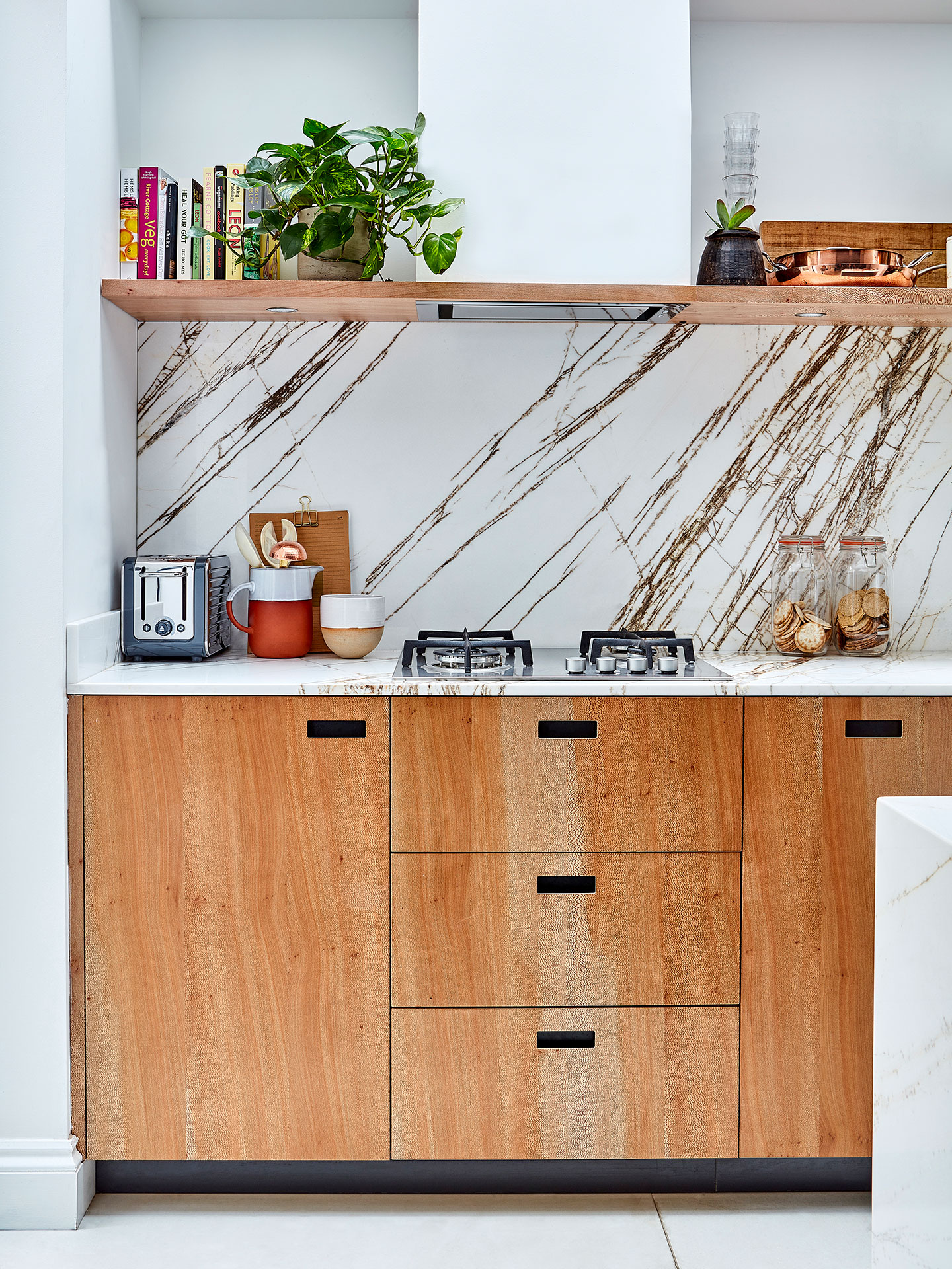 Closeup of a kitchen with wood doors and drawers, a gas hob and a bold marble worktop with matching backsplash.