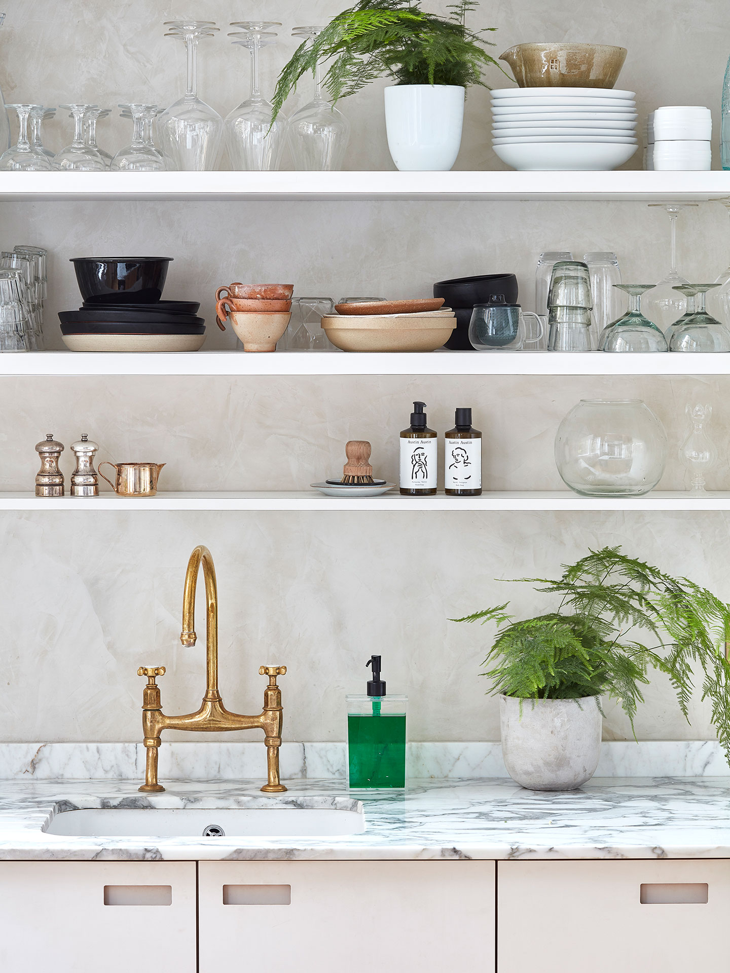 Closeup of a white kitchen with a ceramic sink, brass mixer tap and marble worktops. Above, there are floating shelves.