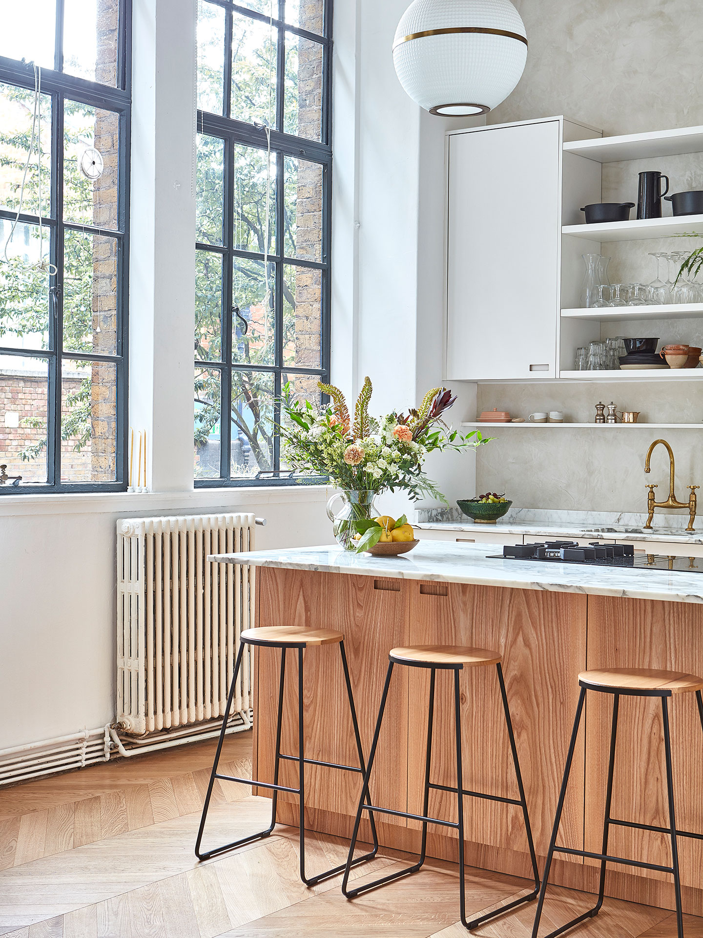 A large kitchen island with wood fronts and marble worktops, behind there is white kitchen cabinetry and crittall windows.