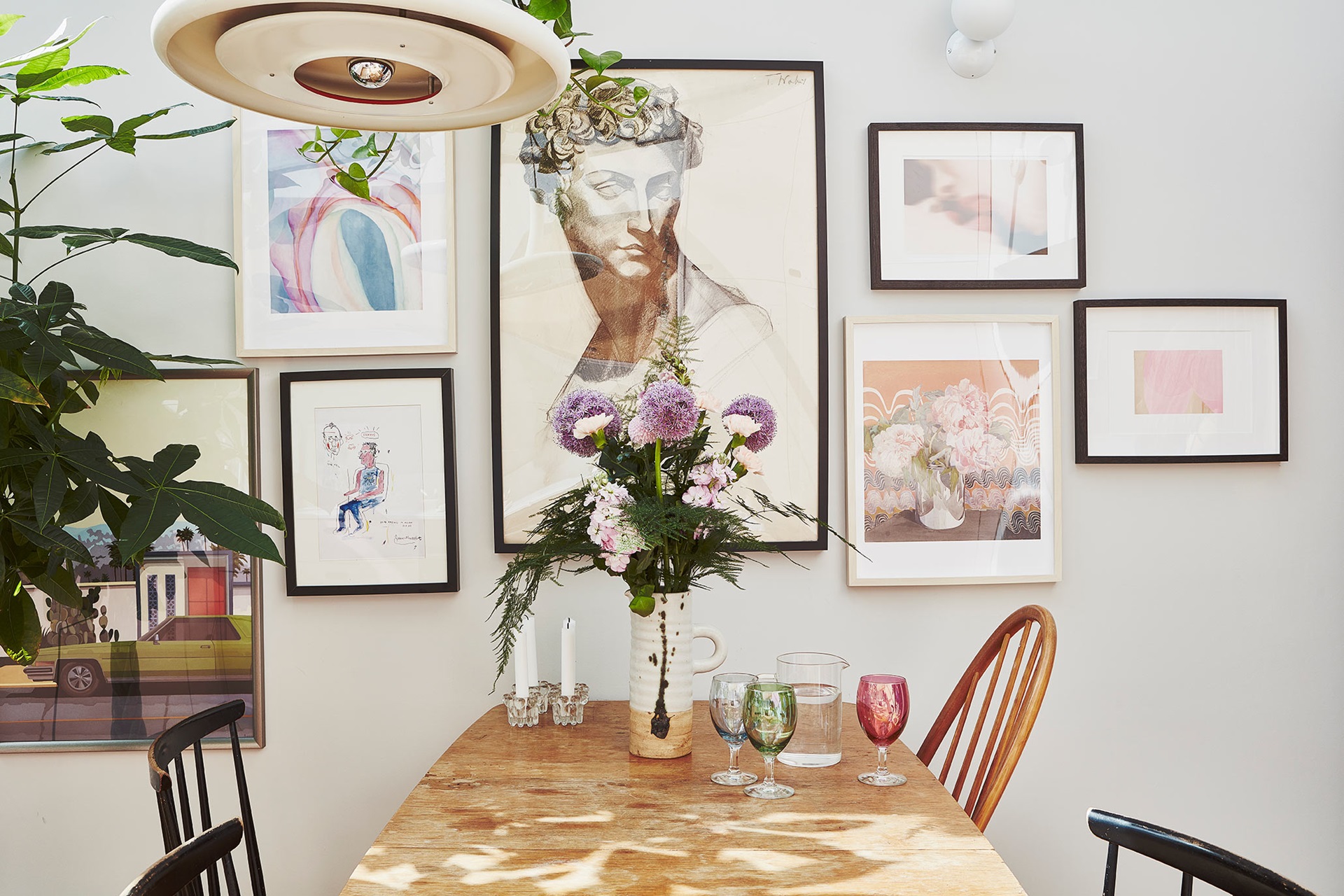 A wood dining table, with colourful glasses and a vase of flowers, sits beneath a gallery wall of paintings and prints.