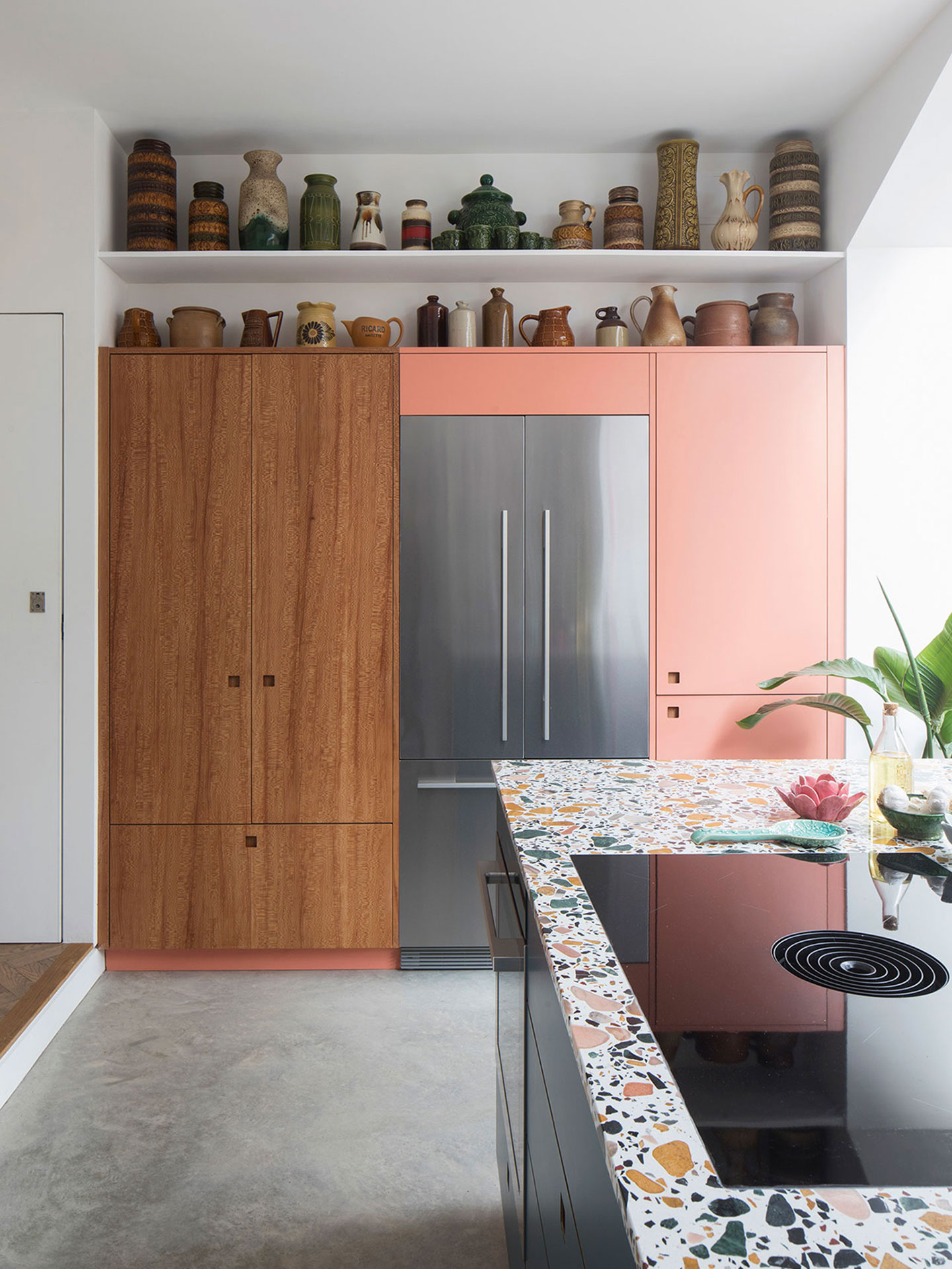 Petrol blue, coral pink and London Plane wood kitchen with a terrazzo topped island and pottery filled shelving.