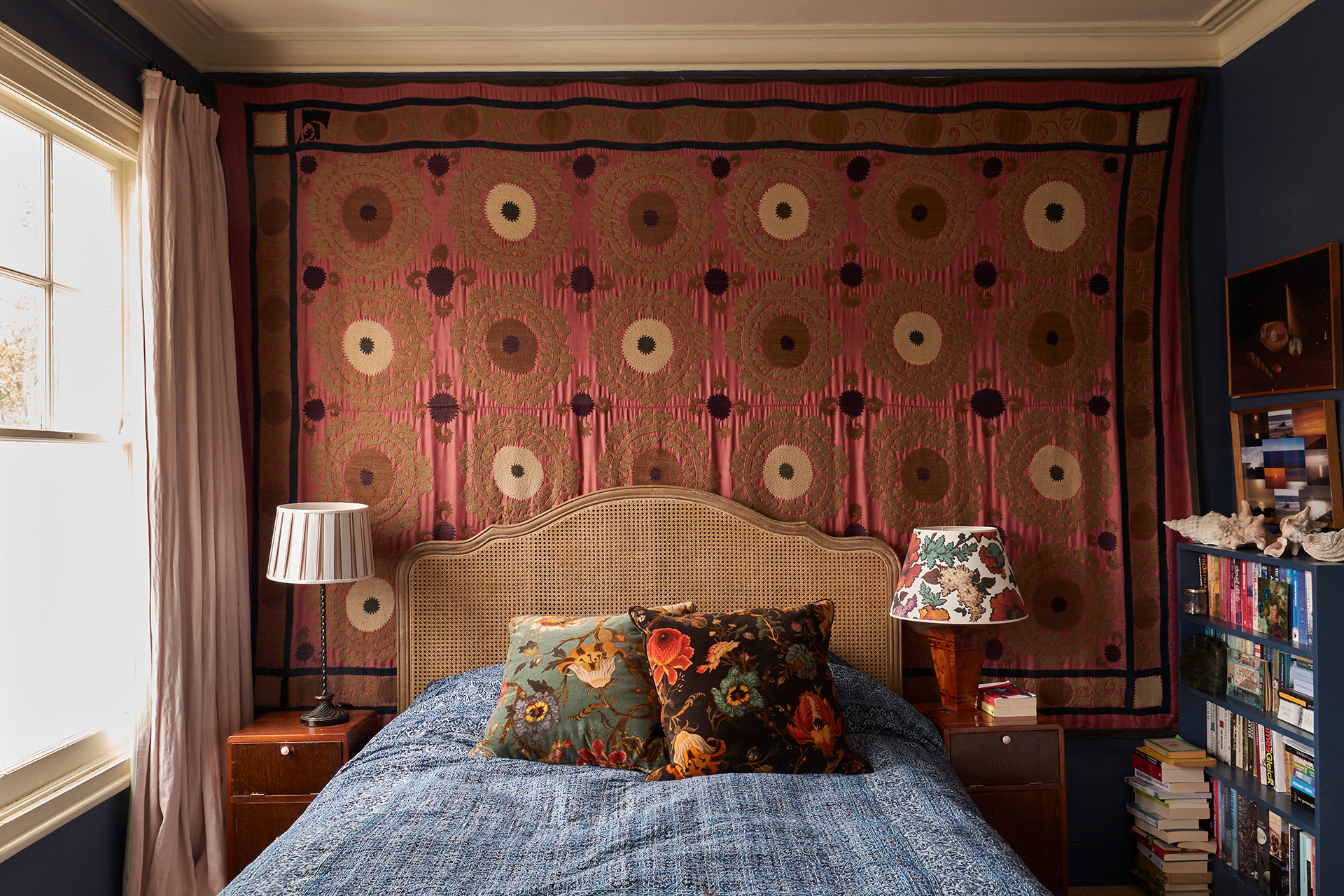 Cosy bedroom with navy blue walls, sash windows and a woven headboard with a pink patterned tapestry hung behind.