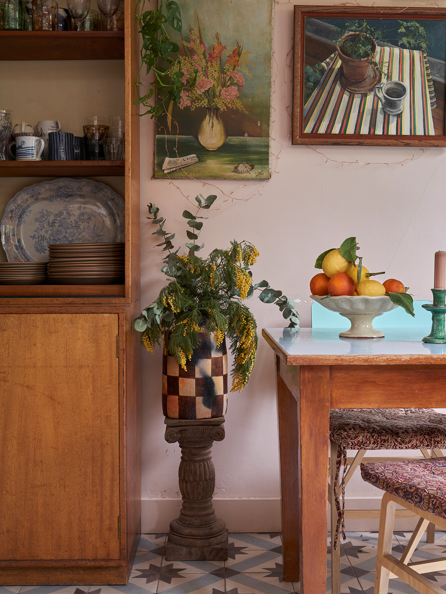 A glass cabinet displaying vintage ceramics sits next to a vase, perched on a plinth, holding eucalyptus and mimosa.