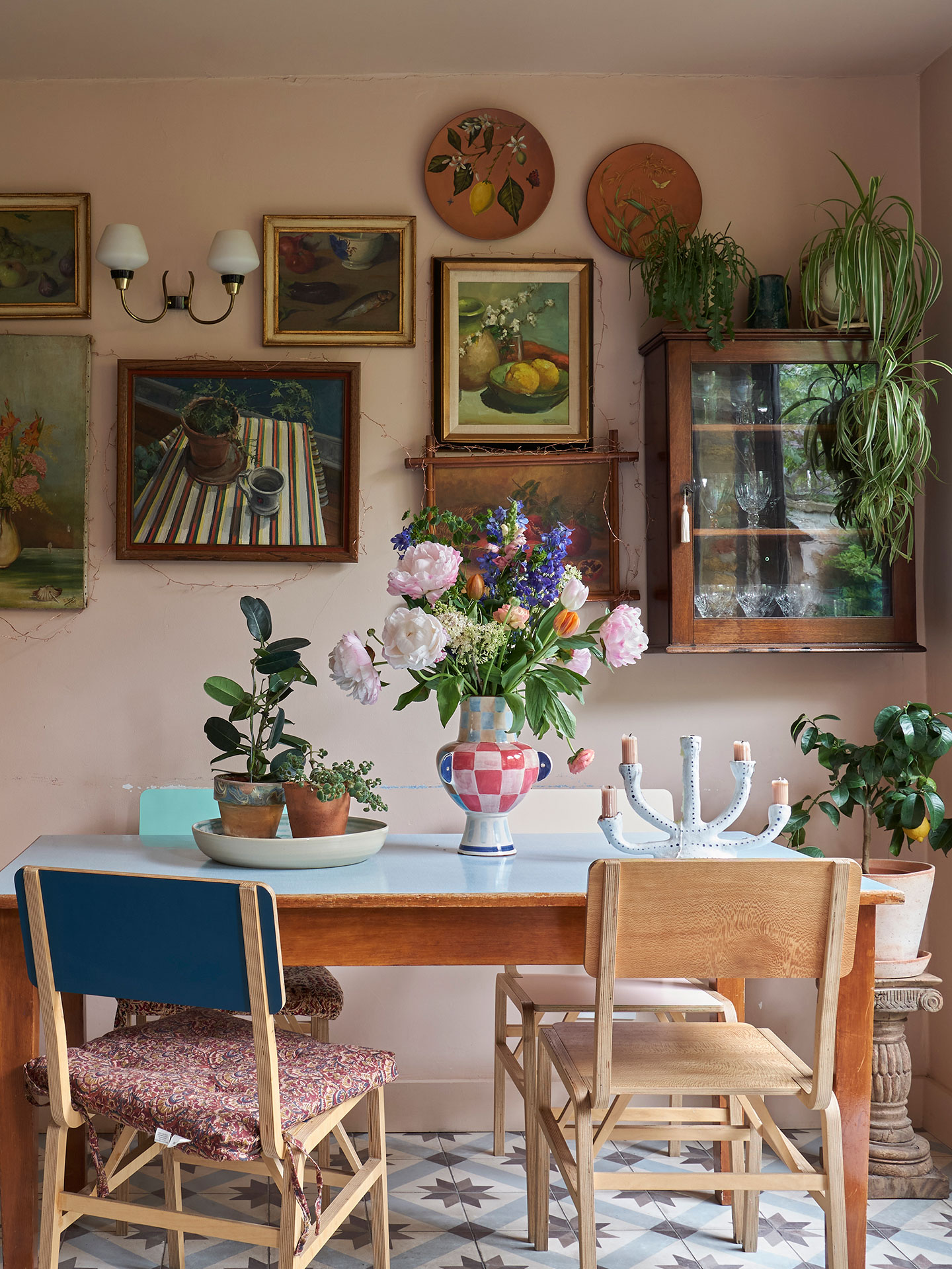 Cosy dining area with a wooden table and mismatched chairs. Paintings and a glass cabinet hang on the pink walls behind.