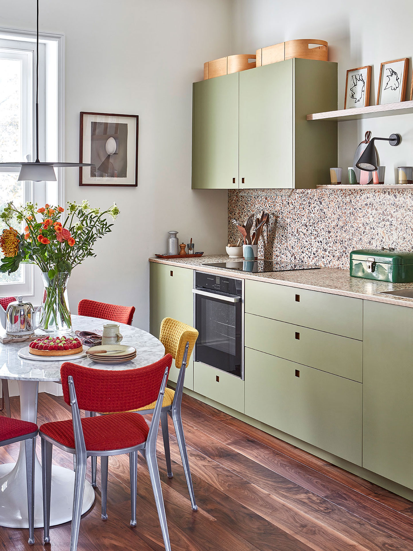 A galley kitchen with green cabinetry and shelving, wood terrazzo worktop and a table with red and yellow chairs.