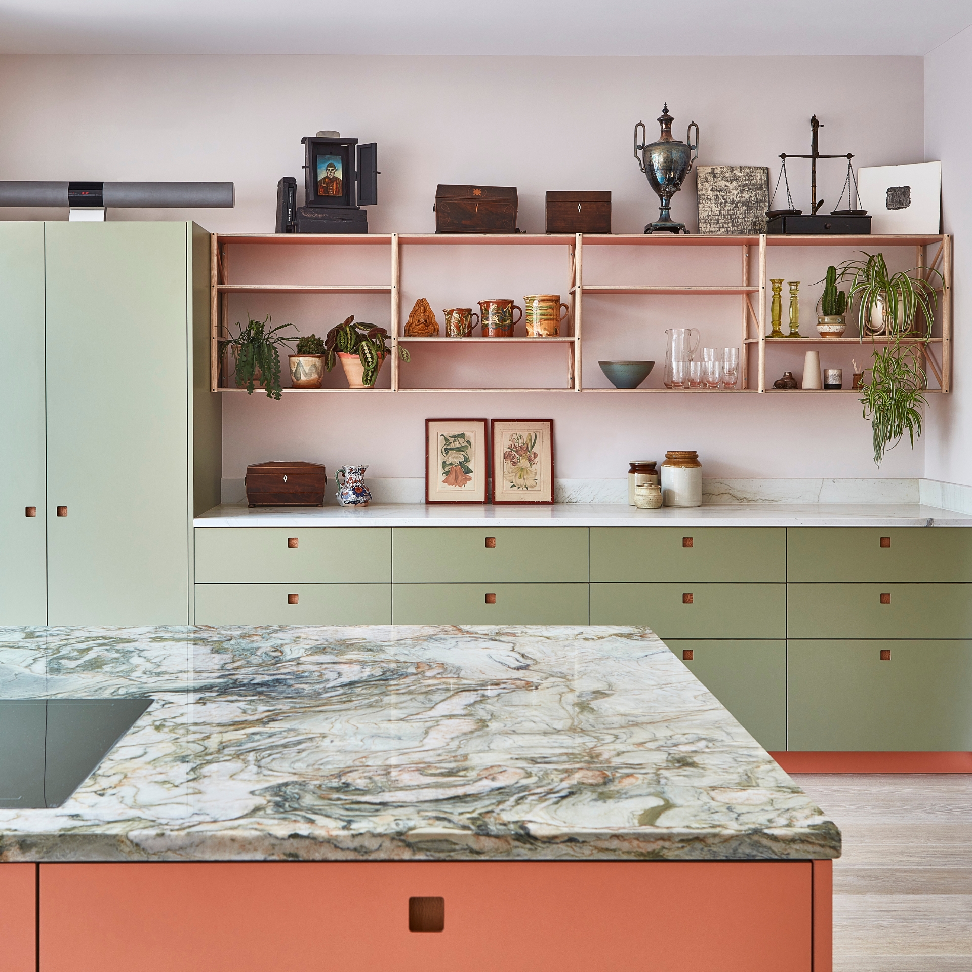 Green kitchen cabinetry with a coral pink island topped with swirling natural stone quartzite and a dining table.