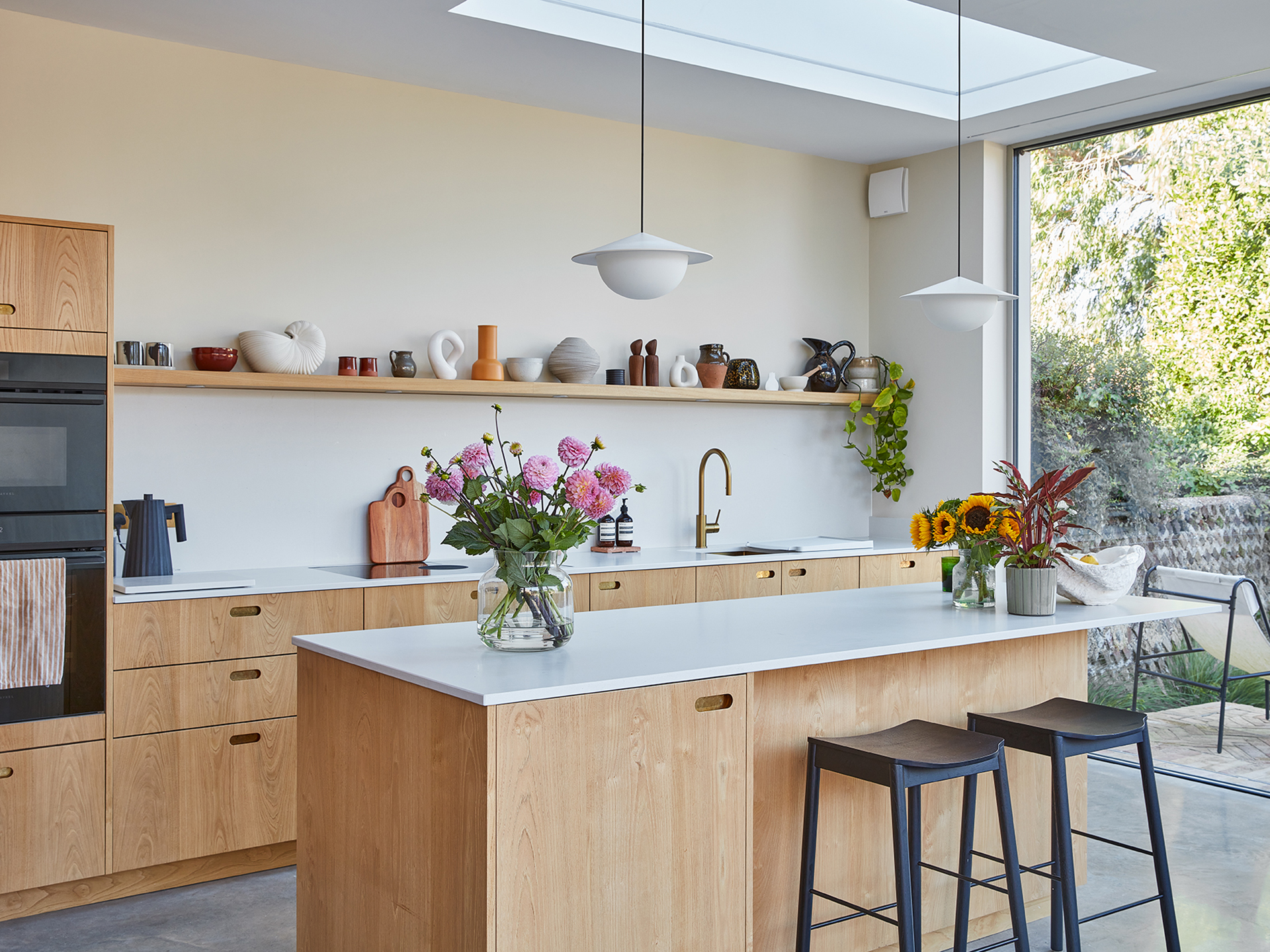 A kitchen in a family home in Brighton with wood cabinetry and brass handles, including an island, larder and shelf and grey concrete floor.