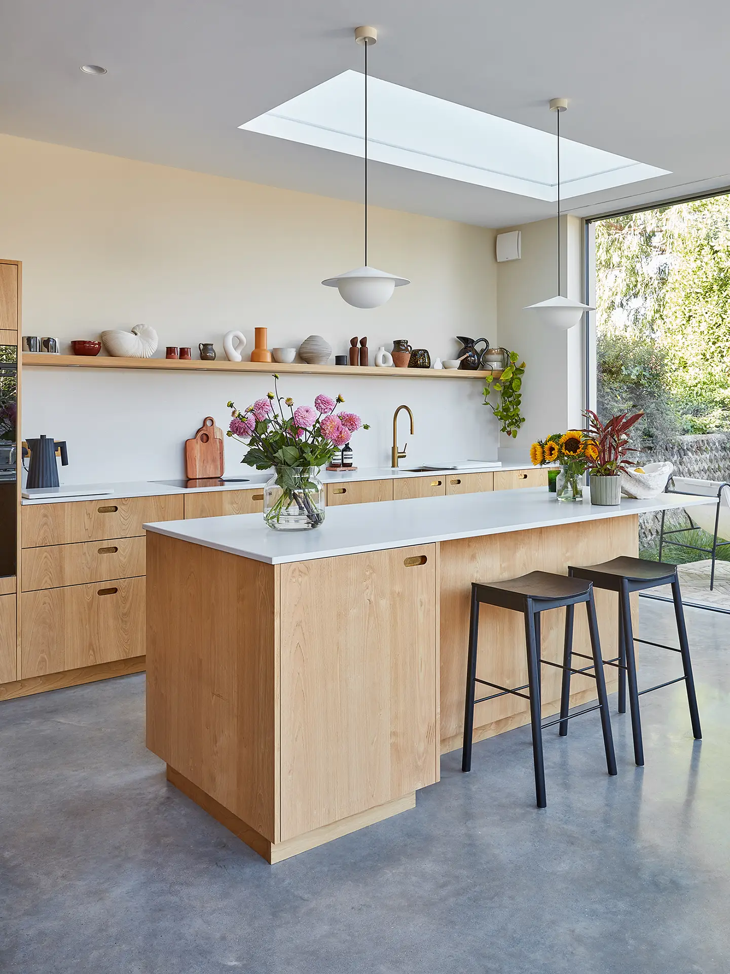 A kitchen in a family home in Brighton with wood cabinetry and brass handles, including an island, larder and shelf and grey concrete floor.