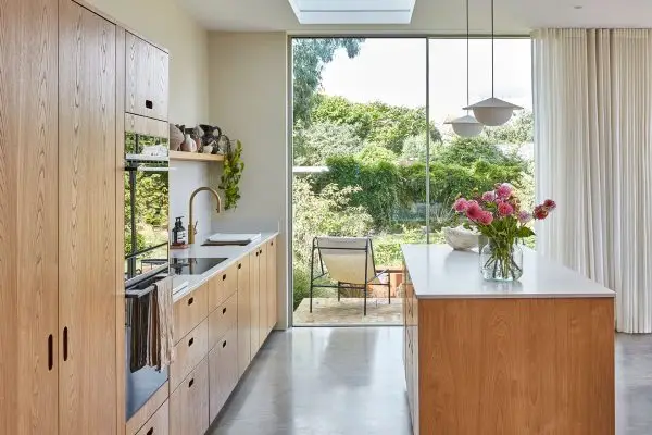 A kitchen in a family home in Brighton with wood cabinetry and brass handles, including an island, larder and shelf and grey concrete floor.