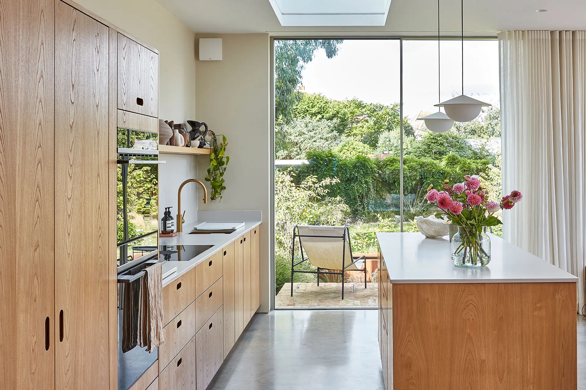 A kitchen in a family home in Brighton with wood cabinetry and brass handles, including an island, larder and shelf and grey concrete floor.