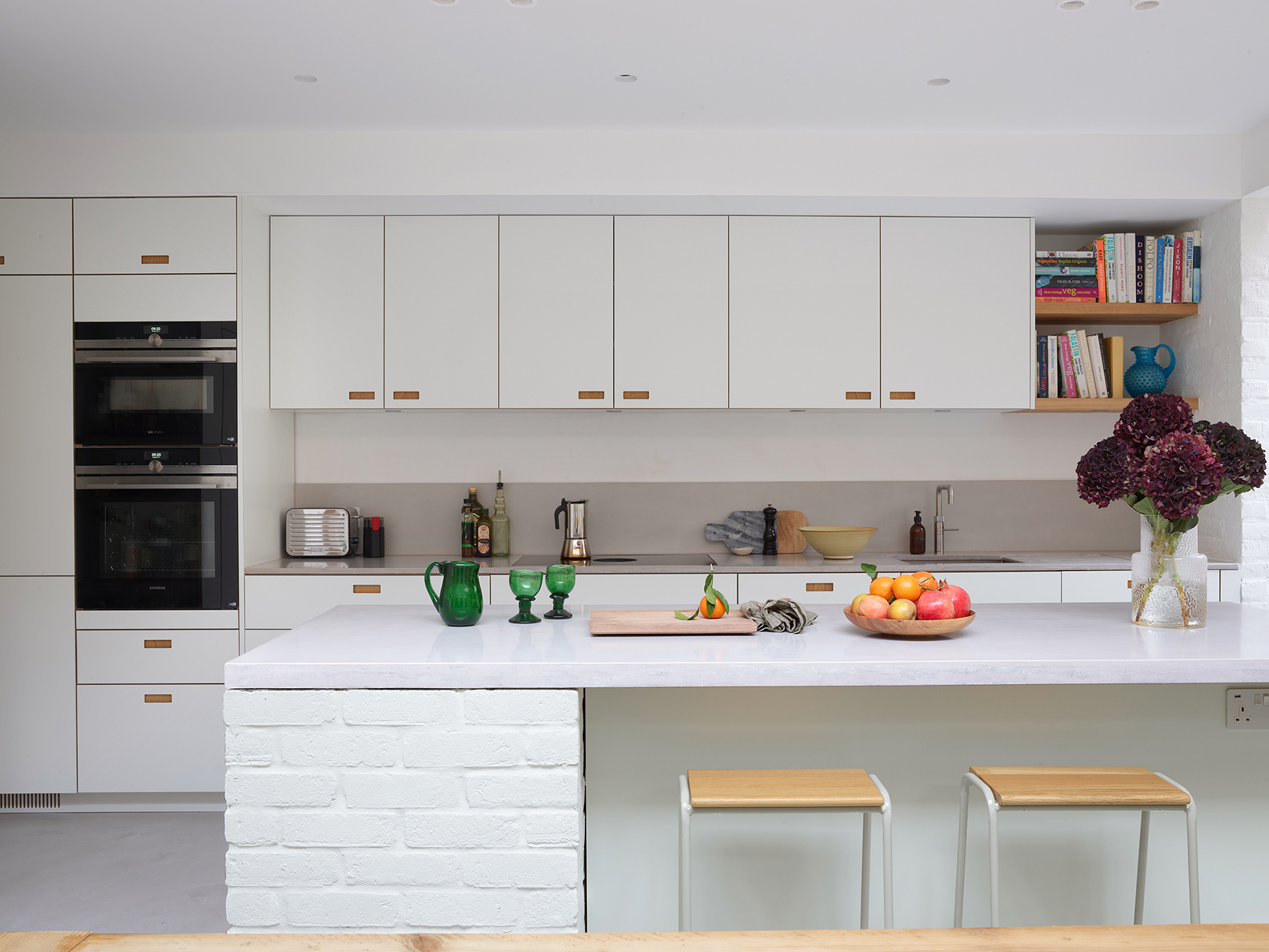 White kitchen in an open plan space with wood beams and exposed painted bricks.