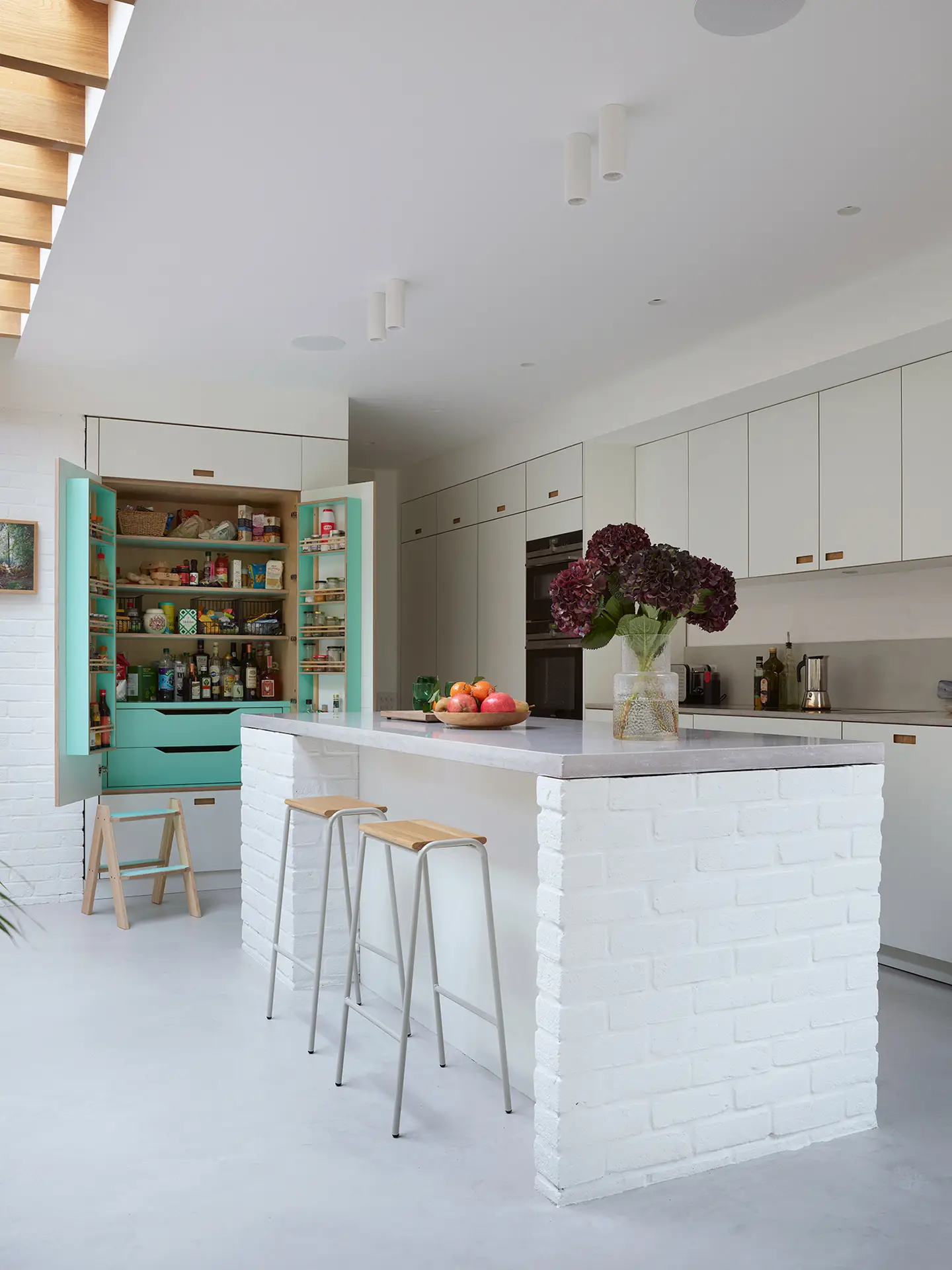 White kitchen in an open plan space with wood beams and exposed painted bricks.