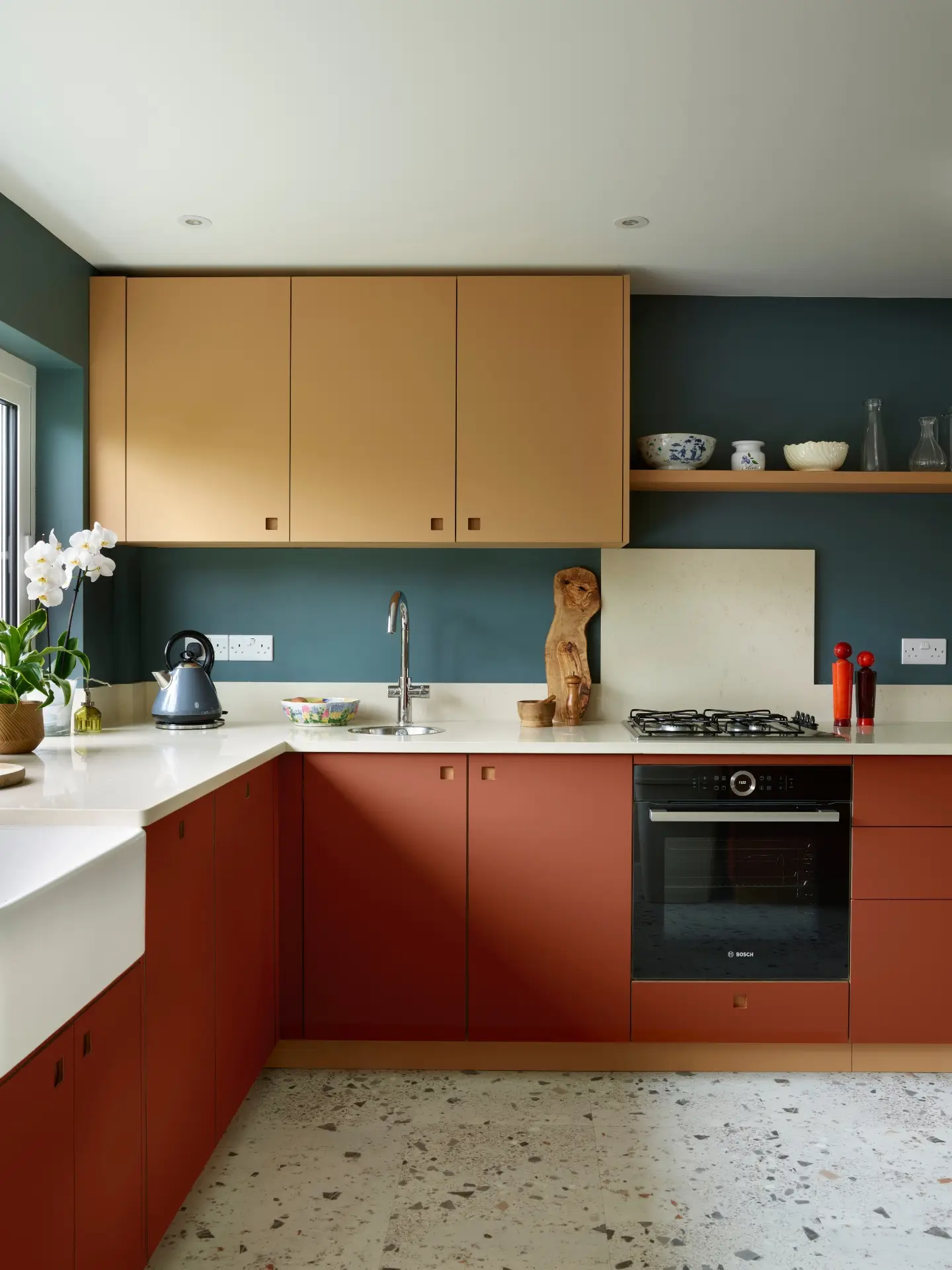 Essex Pluck kitchen with earthy toned ochre and terracotta cabinetry and shelf, white worktops, terrazzo floor and a navy blue wall.