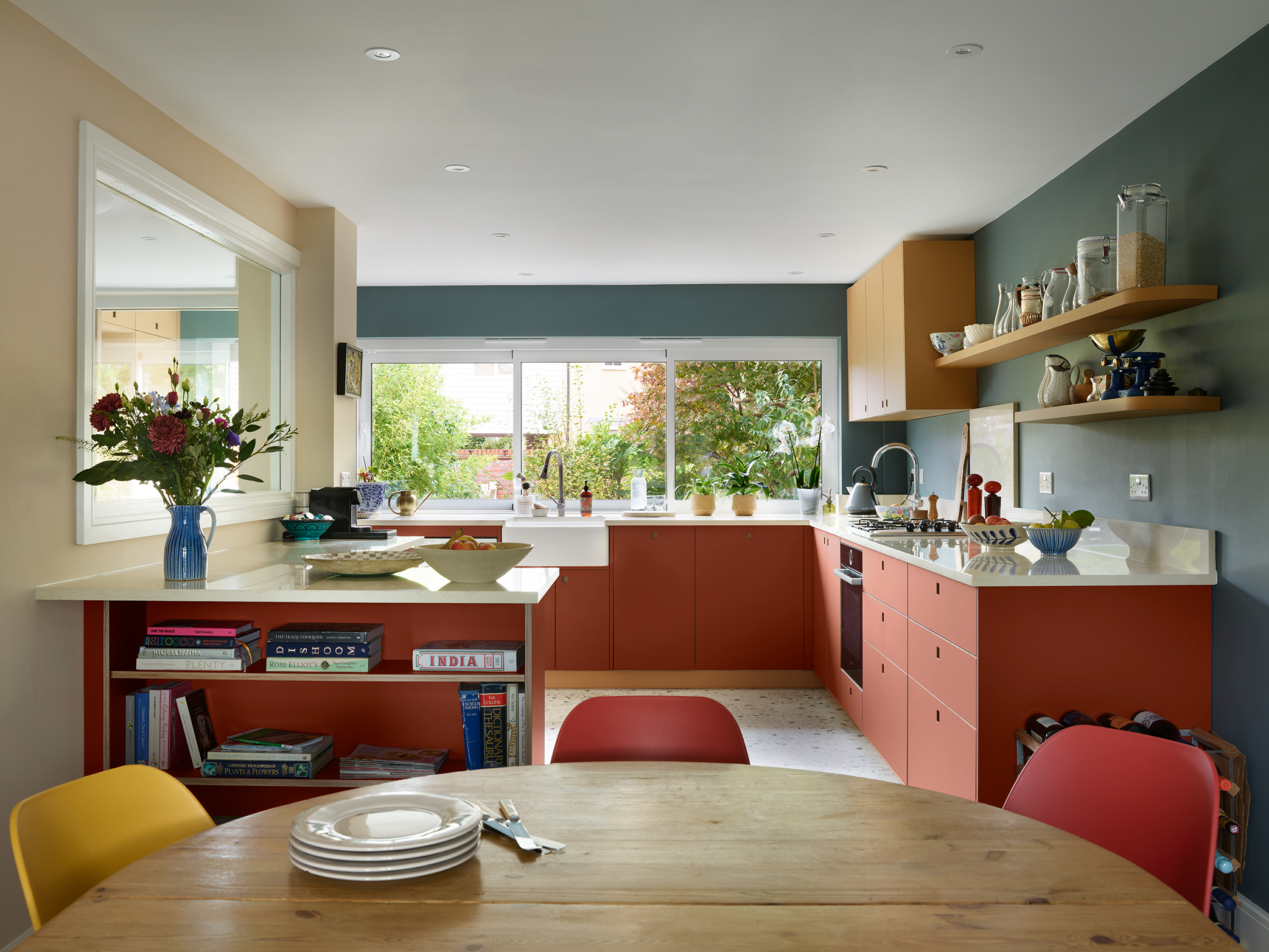 Essex Pluck kitchen with earthy toned ochre and terracotta cabinetry and shelf, white worktops, terrazzo floor and a navy blue wall.