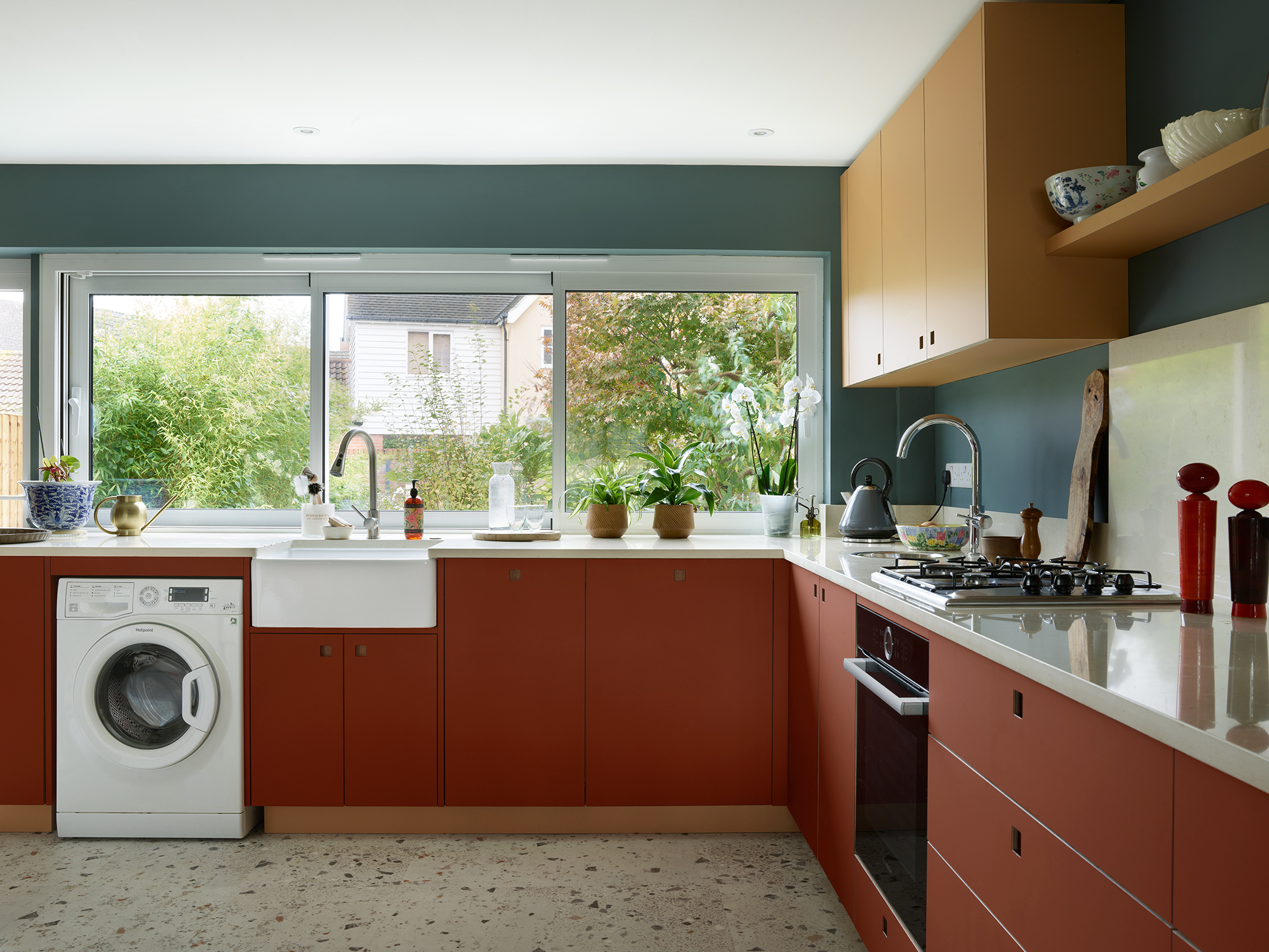 Essex Pluck kitchen with earthy toned ochre and terracotta cabinetry and shelf, white worktops, terrazzo floor and a navy blue wall.