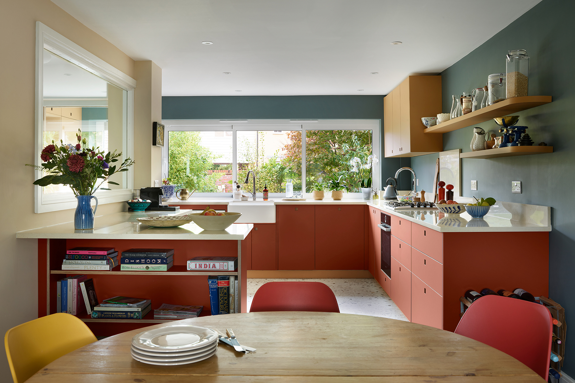 Essex Pluck kitchen with earthy toned ochre and terracotta cabinetry and shelf, white worktops, terrazzo floor and a navy blue wall.