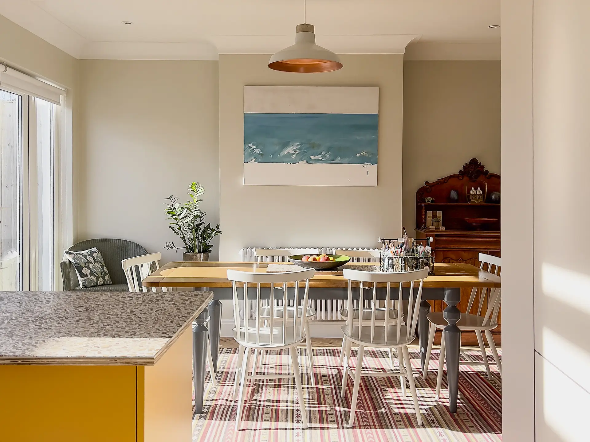 Dining table in a small sunny yellow Pluck kitchen with a peninsula, shelving and glossy tile splashback.