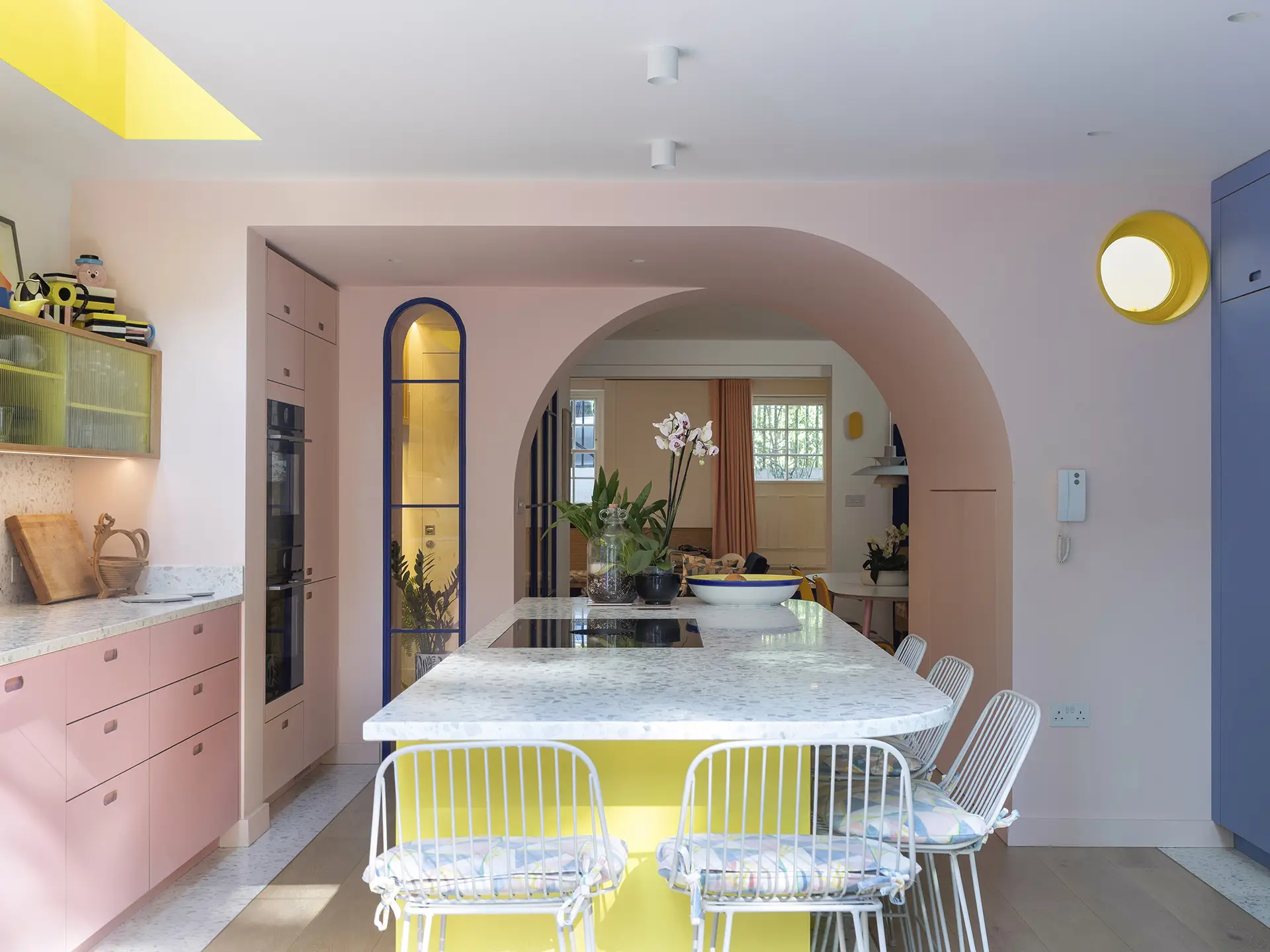 Family kitchen with a playful colour scheme including pink, blue and yellow cabinetry with curved walls, windows and island terrazzo worktop.