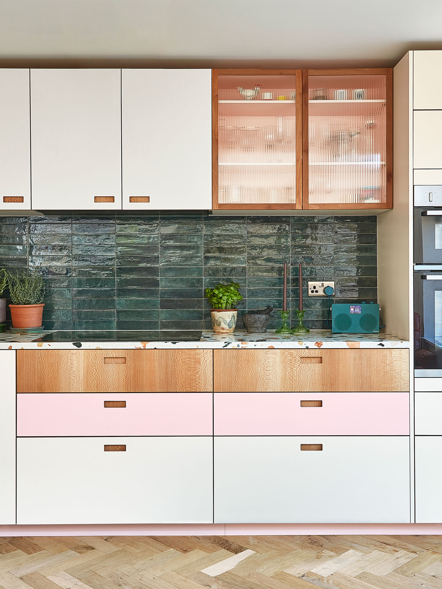 A glass fronted cupboard in a Pluck kitchen with pink, white and London Plane cabinetry, wooden flooring and glossy green tiles.
