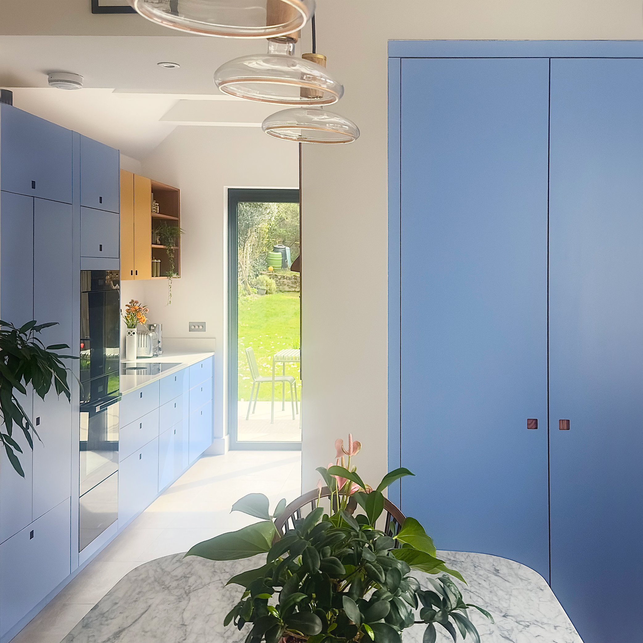 Colourful open plan Pluck kitchen with blue and yellow cabinets and a marble dining table.