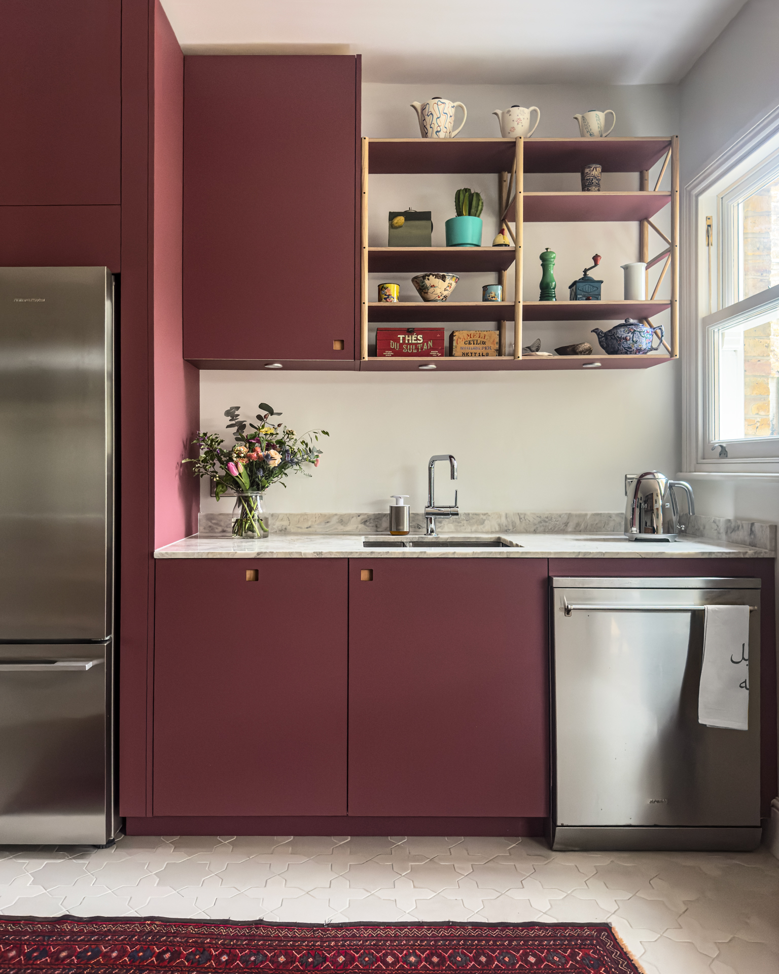 Long narrow kitchen with maroon and navy blue cabinets, open shelving and vintage artwork.