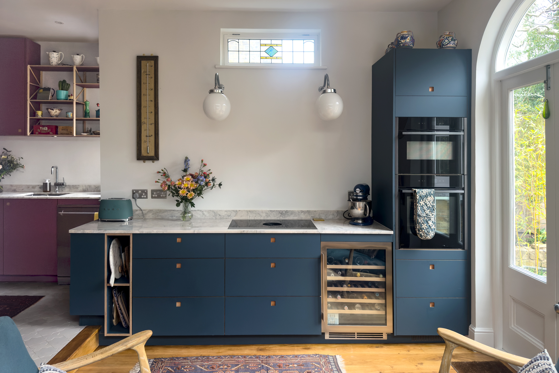A cosy kitchen in Richmond London with pink and navy blue cabinets, stone worktops, vintage art and a wooden floor.