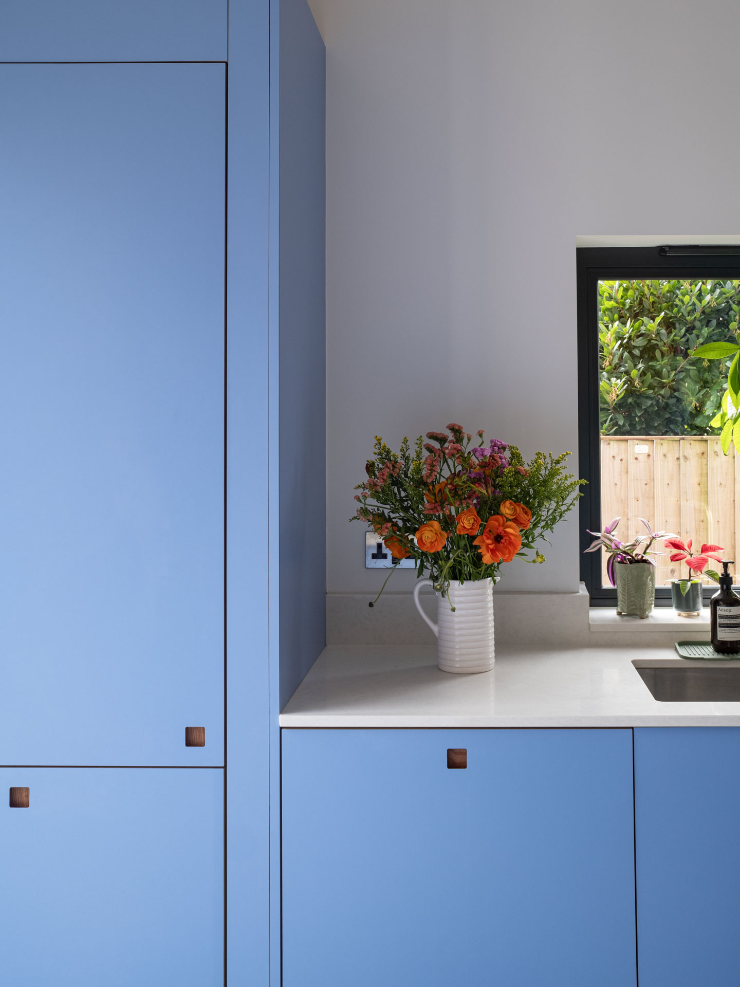 A galley kitchen with blue and yellow cabinetry, a pale pink and elm wood glass cabinet and marble topped dining table.