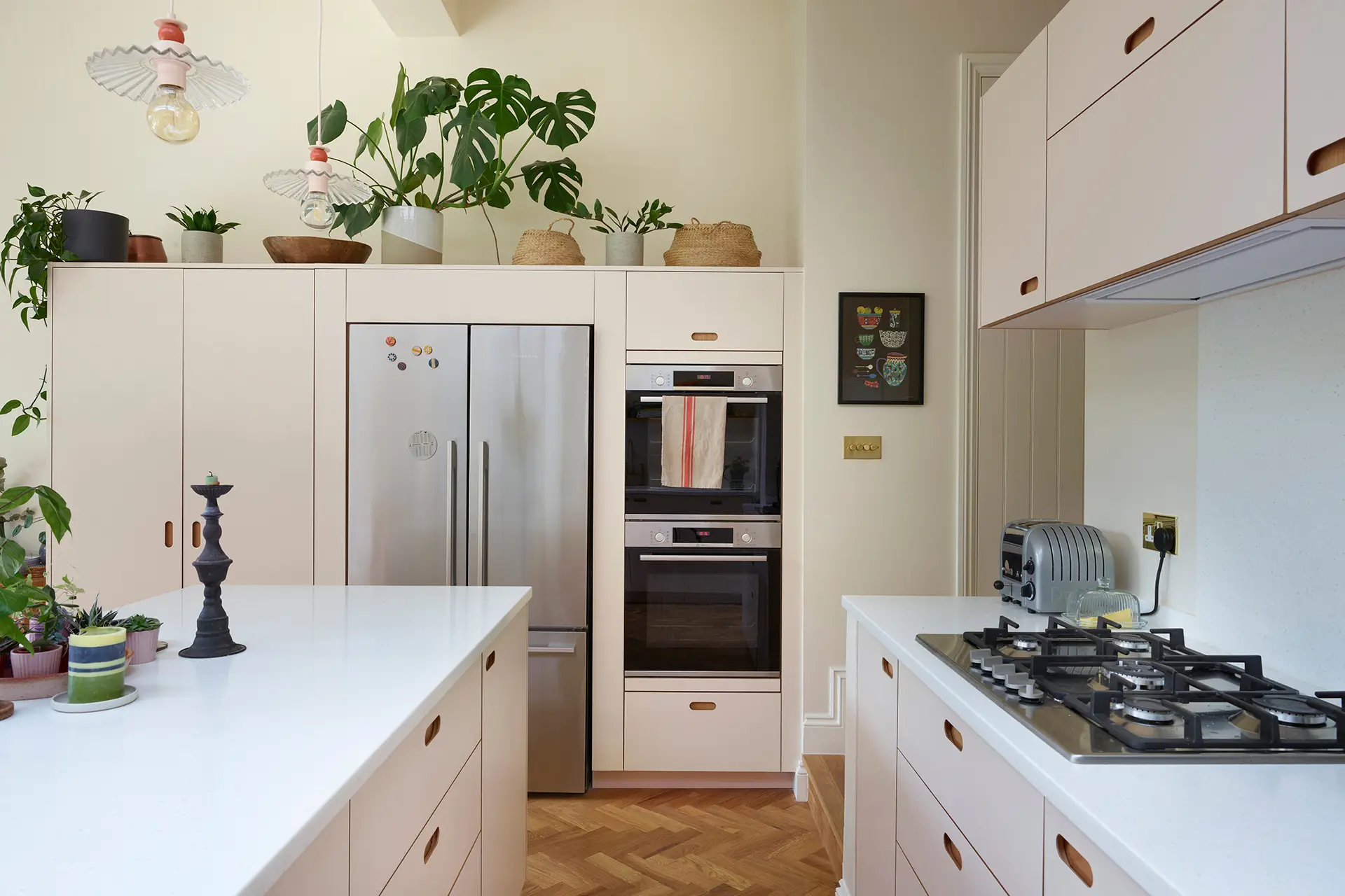 Celebrating neutral hues with beige and soft pink cabinetry in an open plan kitchen and dining area with green glazing and wood floor.