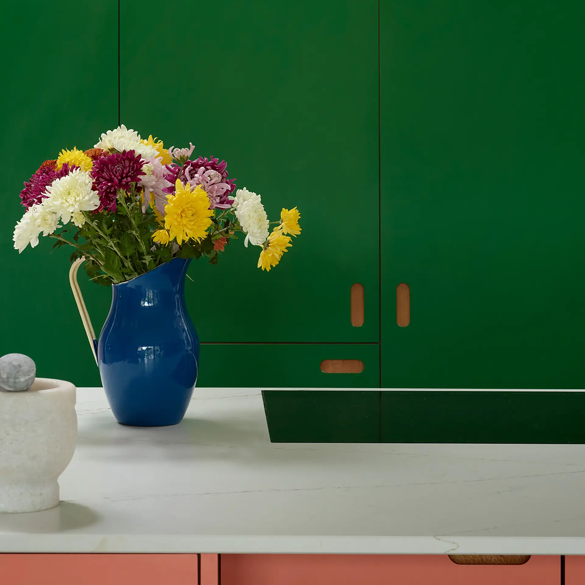 Bold kitchen with pink and green cabinetry, white worktops and a bouquet of flowers.