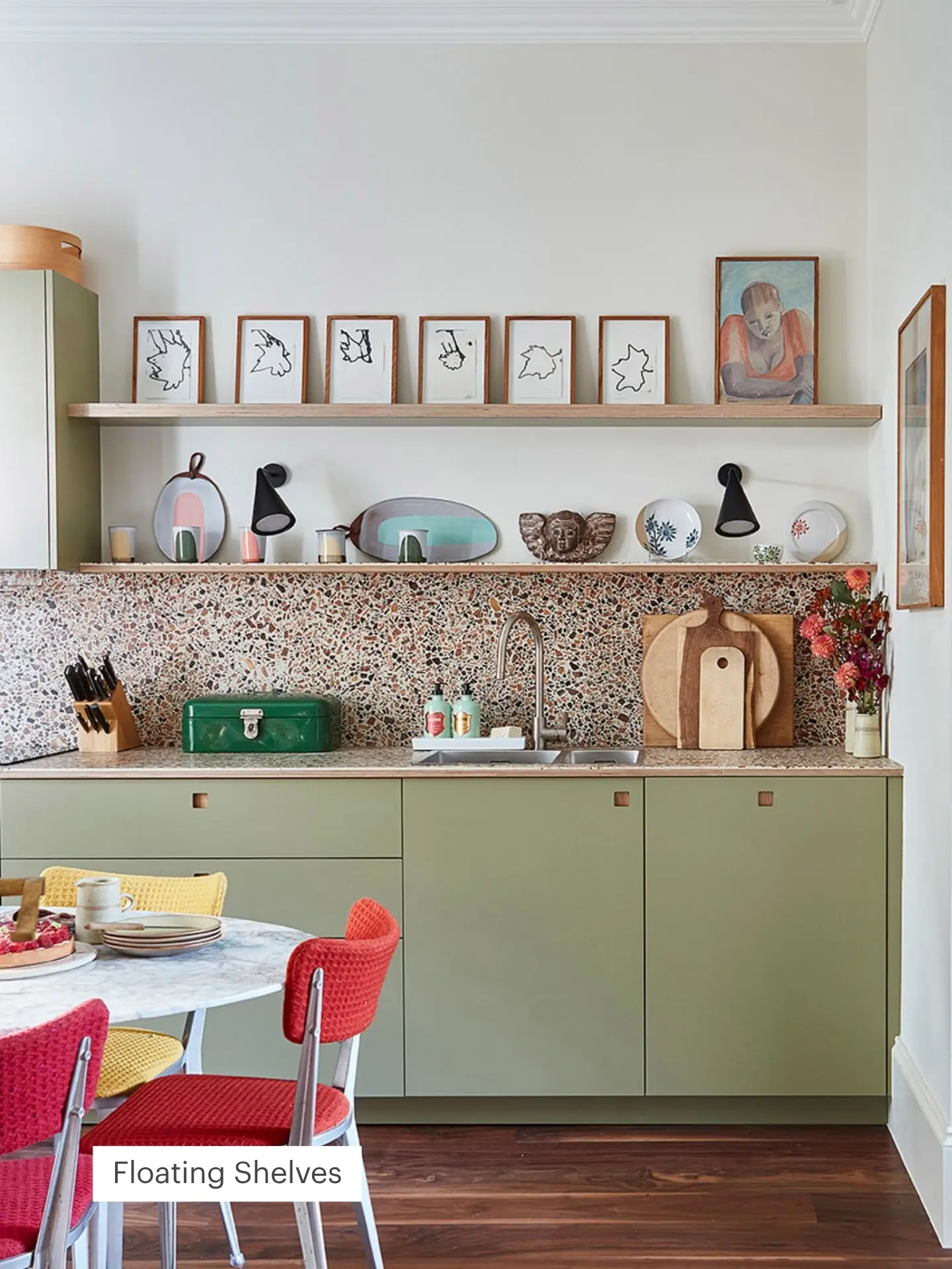 Stylish kitchen with olive green cabinets, terrazzo backsplash, and a wood floating shelf displaying framed art. Bright chairs surround a round table in the foreground.