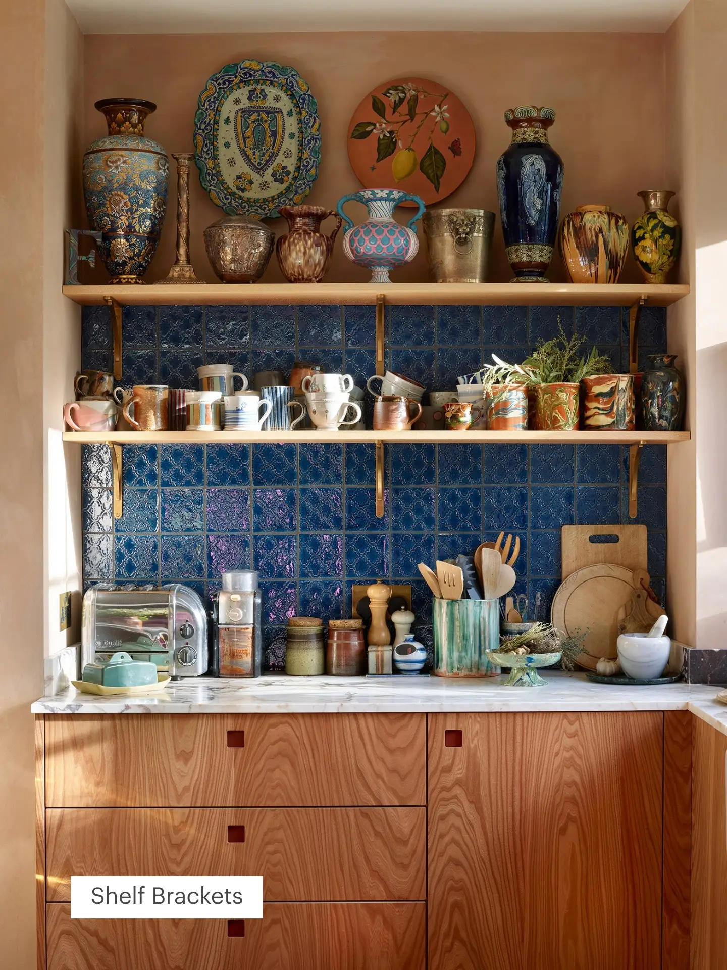 Cosy kitchen with blue tile backsplash, wooden shelves holding ceramic mugs and vases, and a marble counter with jars, toaster, and kitchen tools.