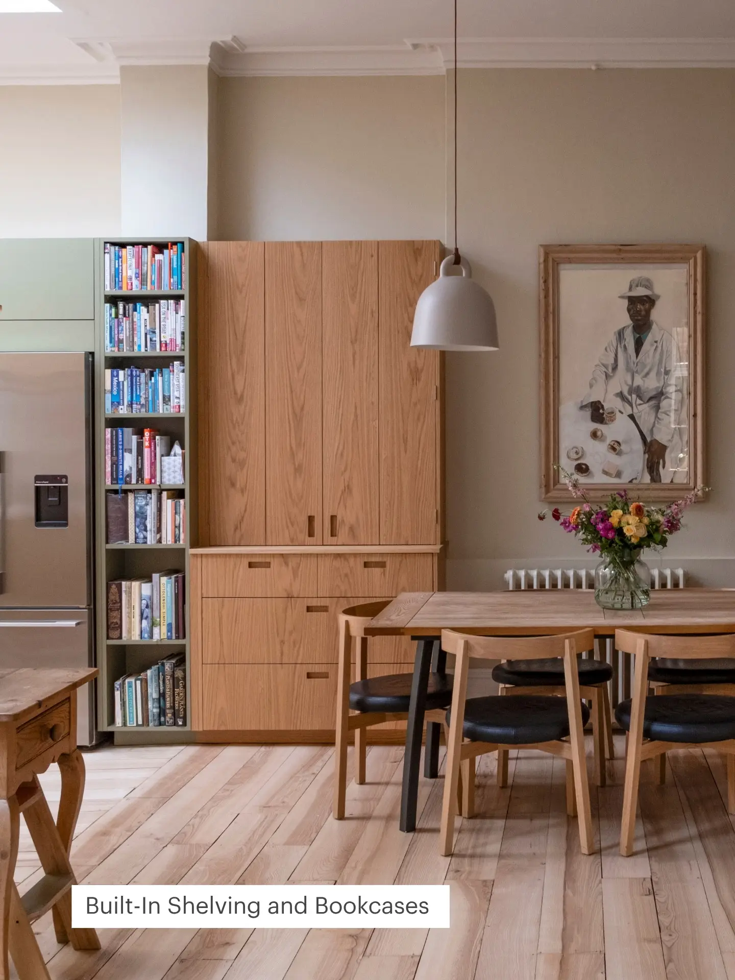 A cosy open plan kitchen with wooden furniture. Built-in shelves filled with books sit beside a fridge. A painting hangs above a table with a flower vase.