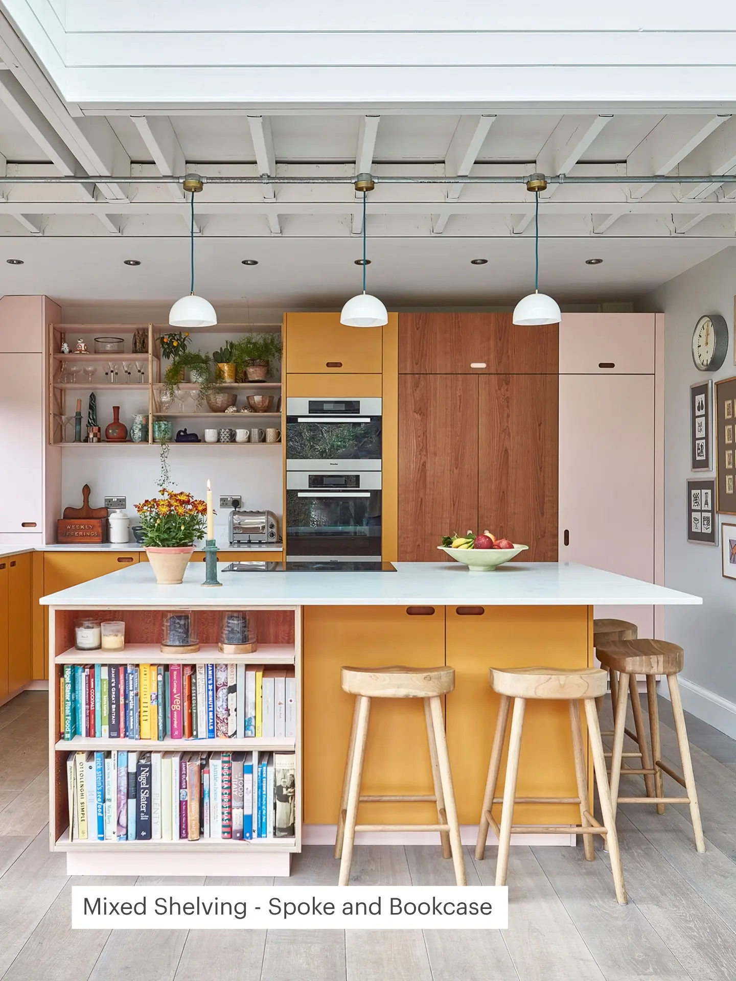 Modern kitchen with pastel pink and mustard yellow cabinets, open shelving with plants, books, and decor. A large island features wooden stools and a fruit bowl.