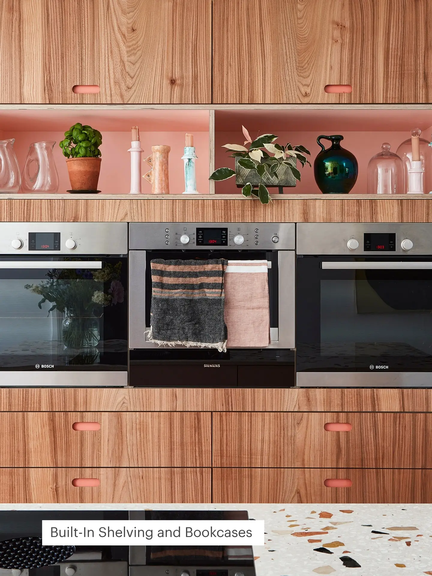 Warm kitchen with wood cabinets, modern appliances and pink shelves dressed with lush plants and colourful ceramics.