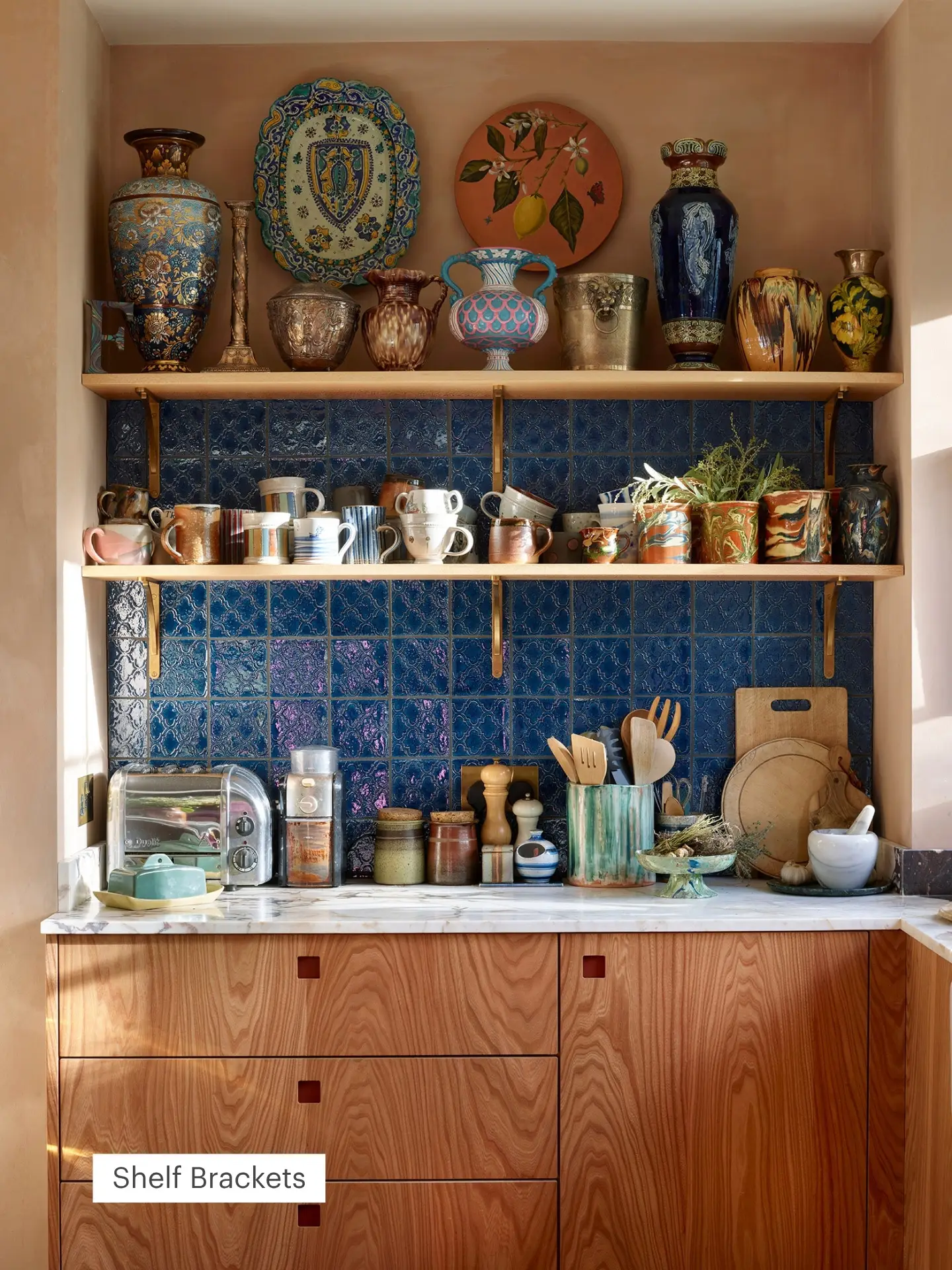 Cosy kitchen with blue tile backsplash, wooden shelves holding ceramic mugs and vases, and a marble counter with jars, toaster, and kitchen tools.