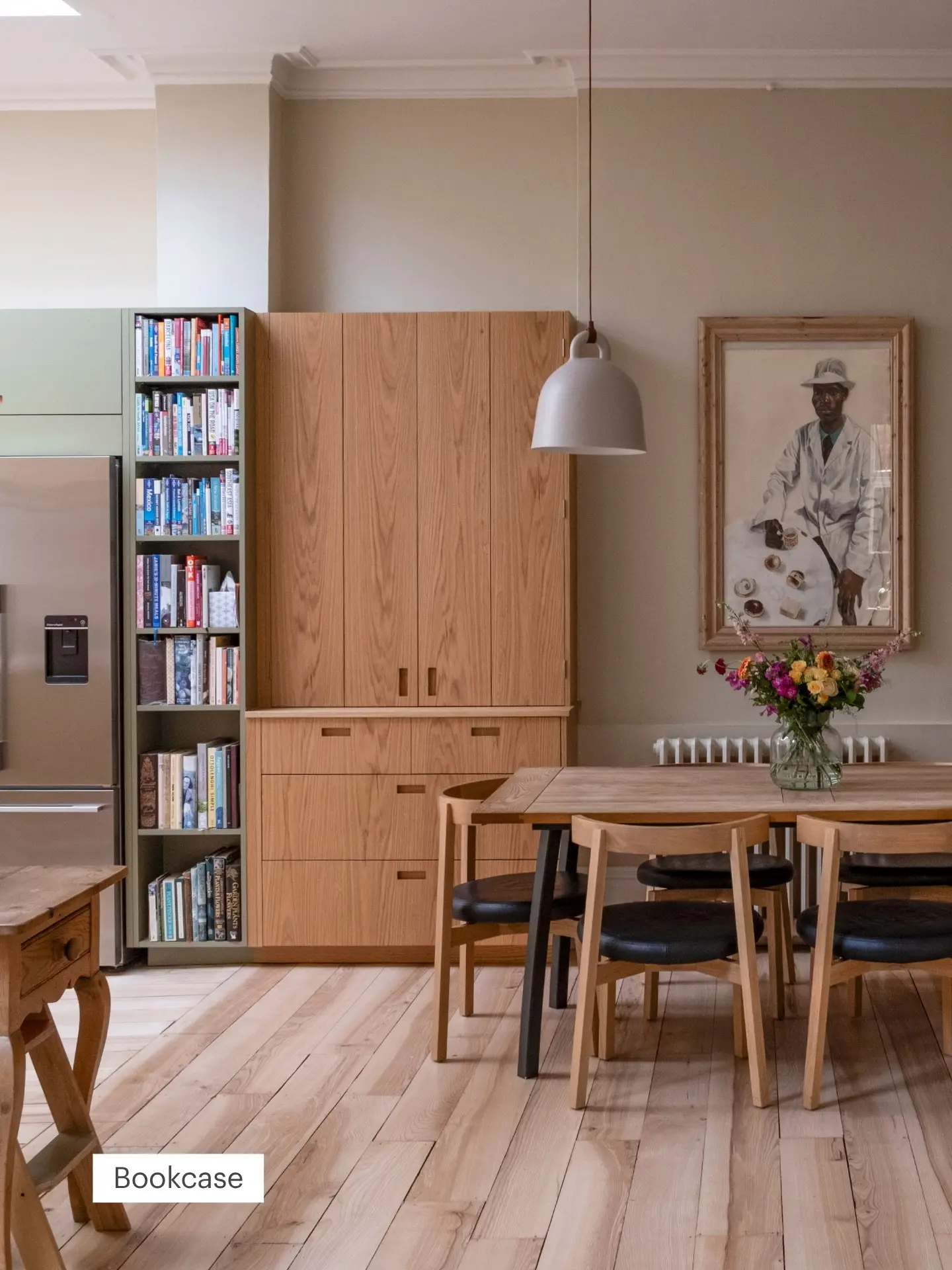 A cosy open plan kitchen with wooden furniture. Built-in shelves filled with books sit beside a fridge. A painting hangs above a table with a flower vase.