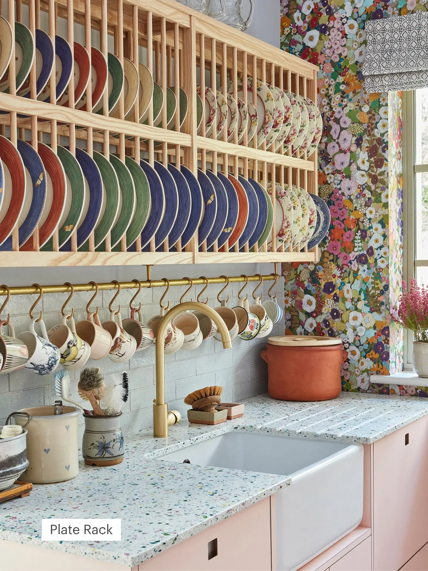 A cosy kitchen with colourful plates neatly arranged in a wooden plate rack, hanging mugs, terrazzo countertop, floral wallpaper, and a farmhouse sink.
