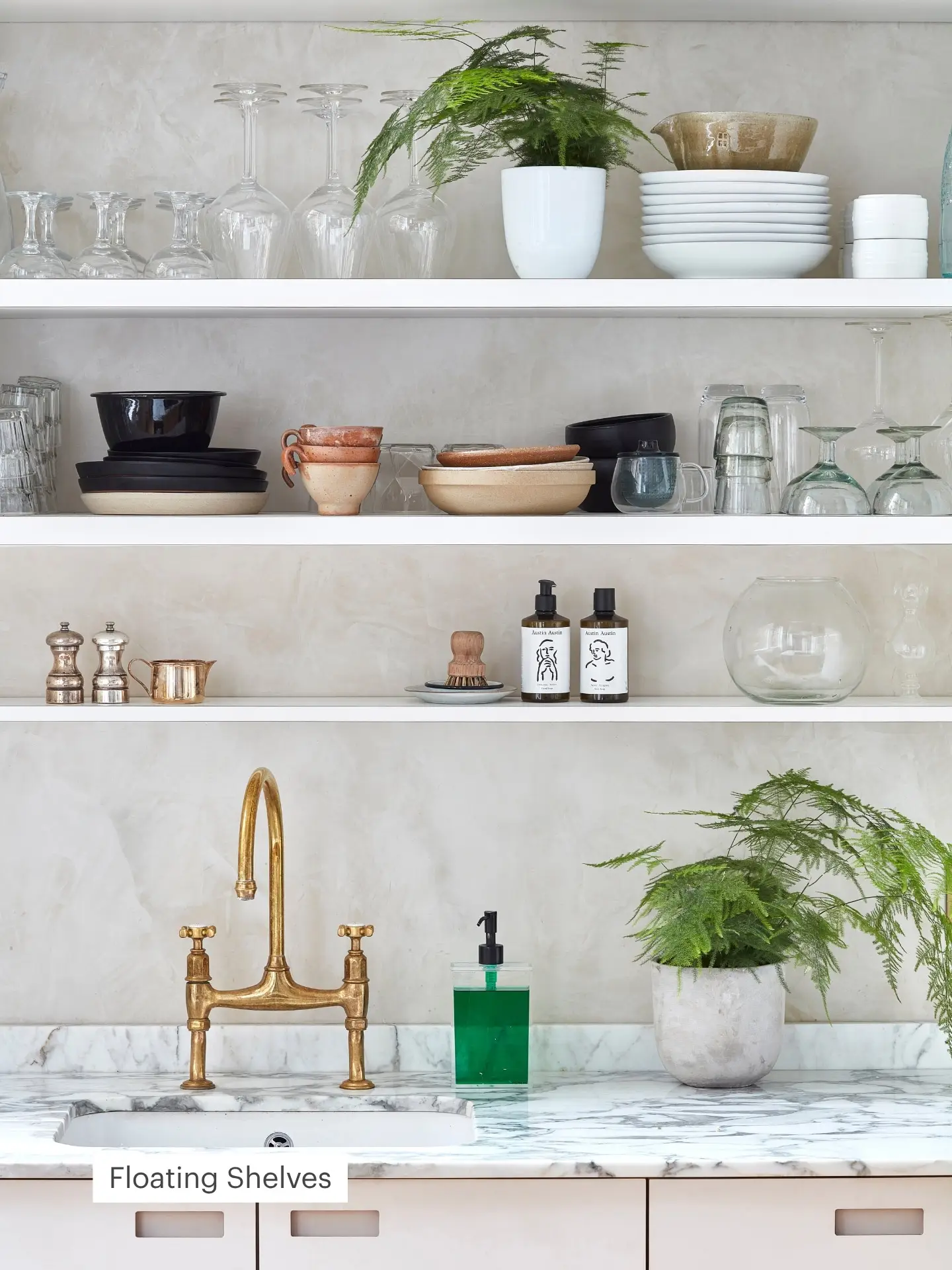 Closeup of a Pluck kitchen with marble worktops, brass faucet, three floating shelves holding glassware, dishes, a potted plant, and bottles.