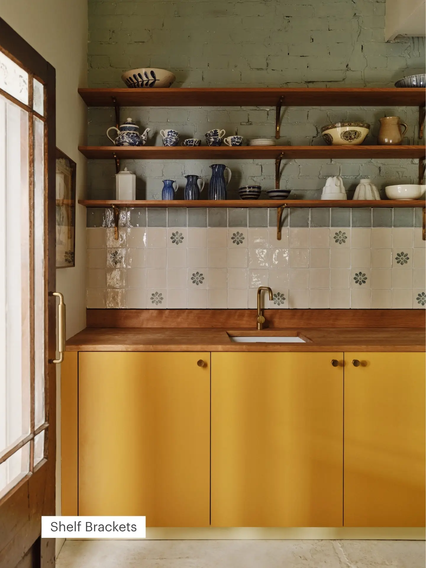 A Pluck kitchen with open wooden shelves displaying blue and white ceramics, yellow cabinets, a wooden countertop, and floral white tile backsplash.