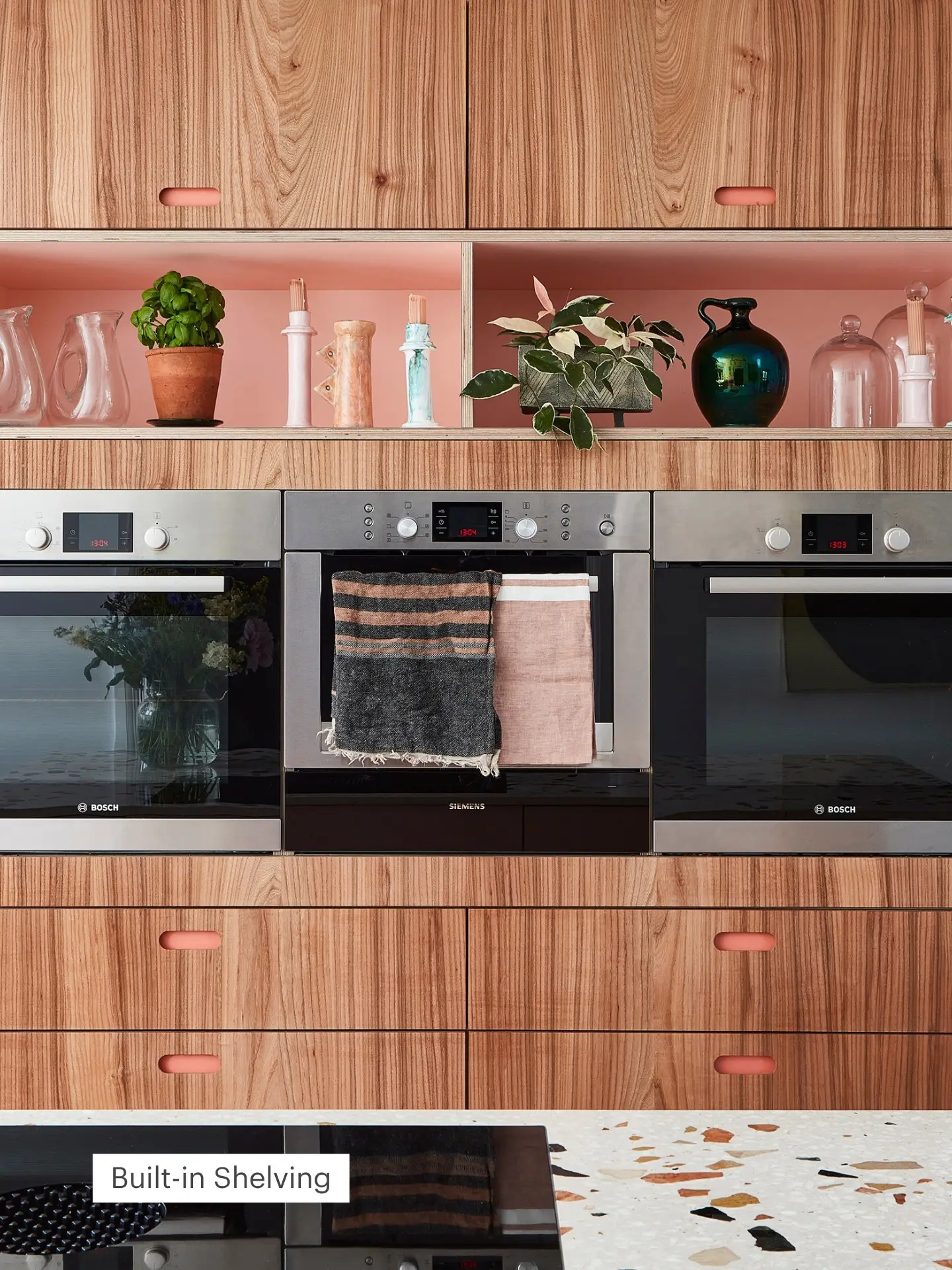 Warm kitchen with wood cabinets, modern appliances and pink shelves dressed with lush plants and colourful ceramics.