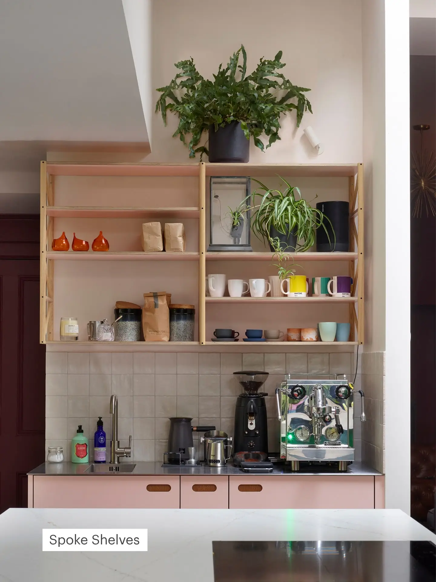 Coffee station in a pluck kitchen with pink cabinetry, stainless steel worktops, pink spoke shelves brimming with mugs and a large coffee machine.