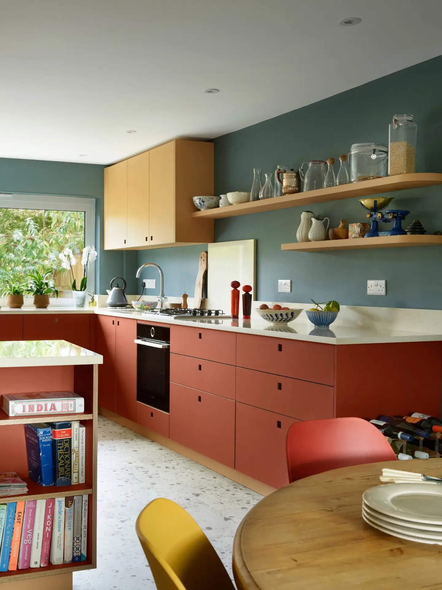Modern kitchen with warm tones featuring terracotta cabinets, open wooden shelves with various dishes, a bookcase, and a round table with colourful chairs.