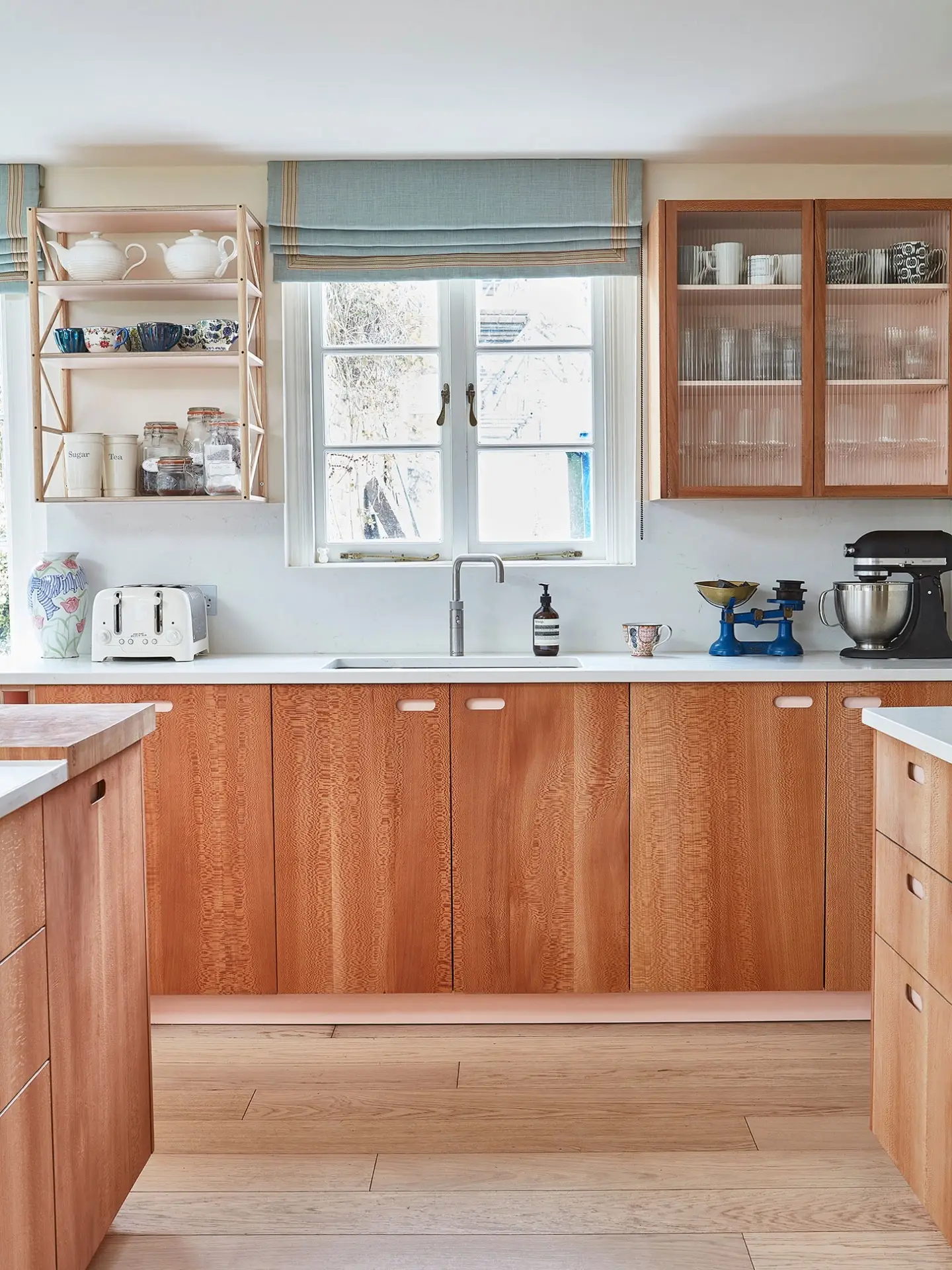 A bright kitchen with wooden cabinets and a white worktop featuring a silver tap, toaster and kitchen aid mixer. Open shelves hold jars and tea sets.