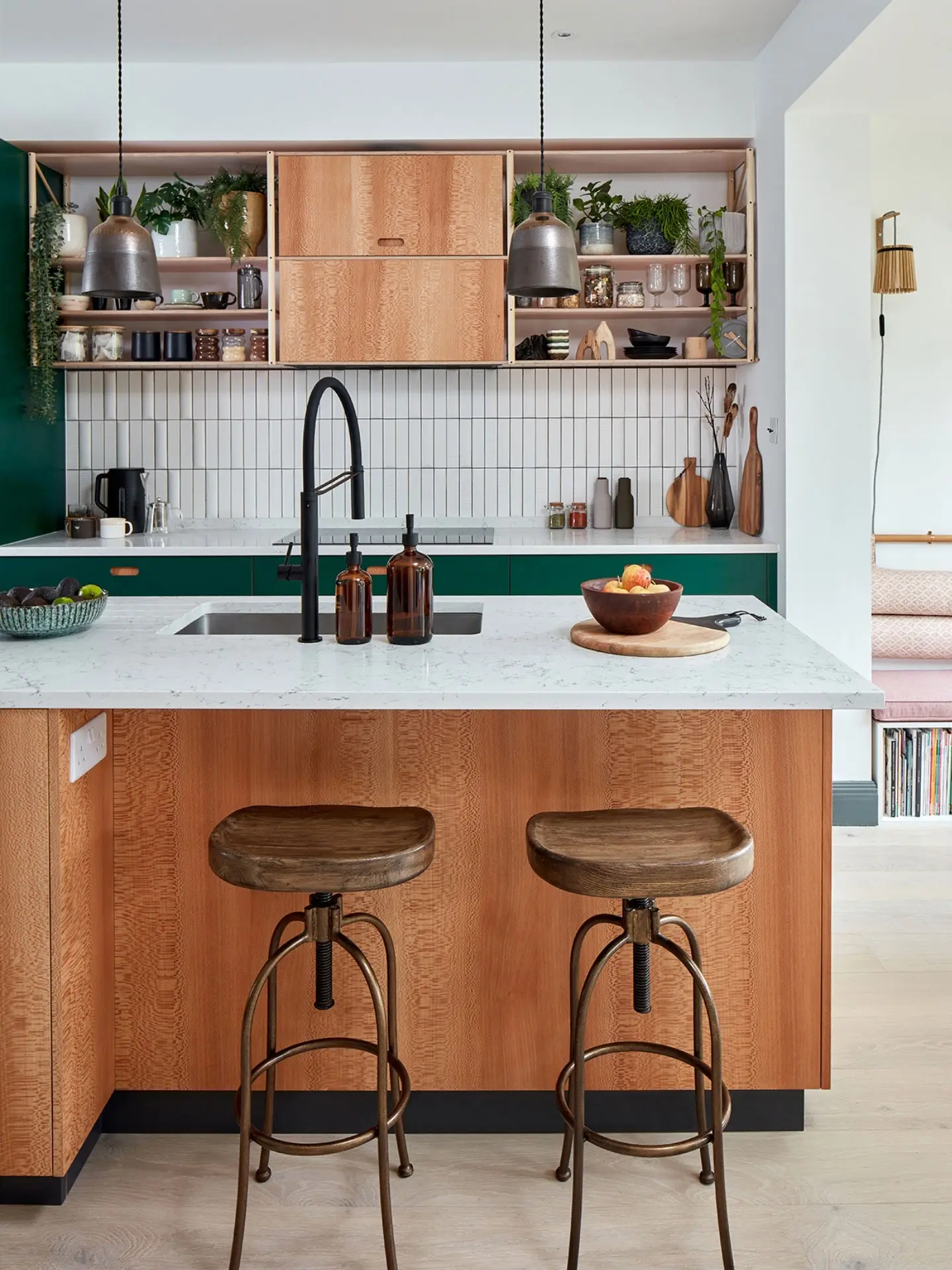 A modern kitchen with marble worktops, a large kitchen island with wooden stools and shelves on the wall behind displays plants and ceramics.