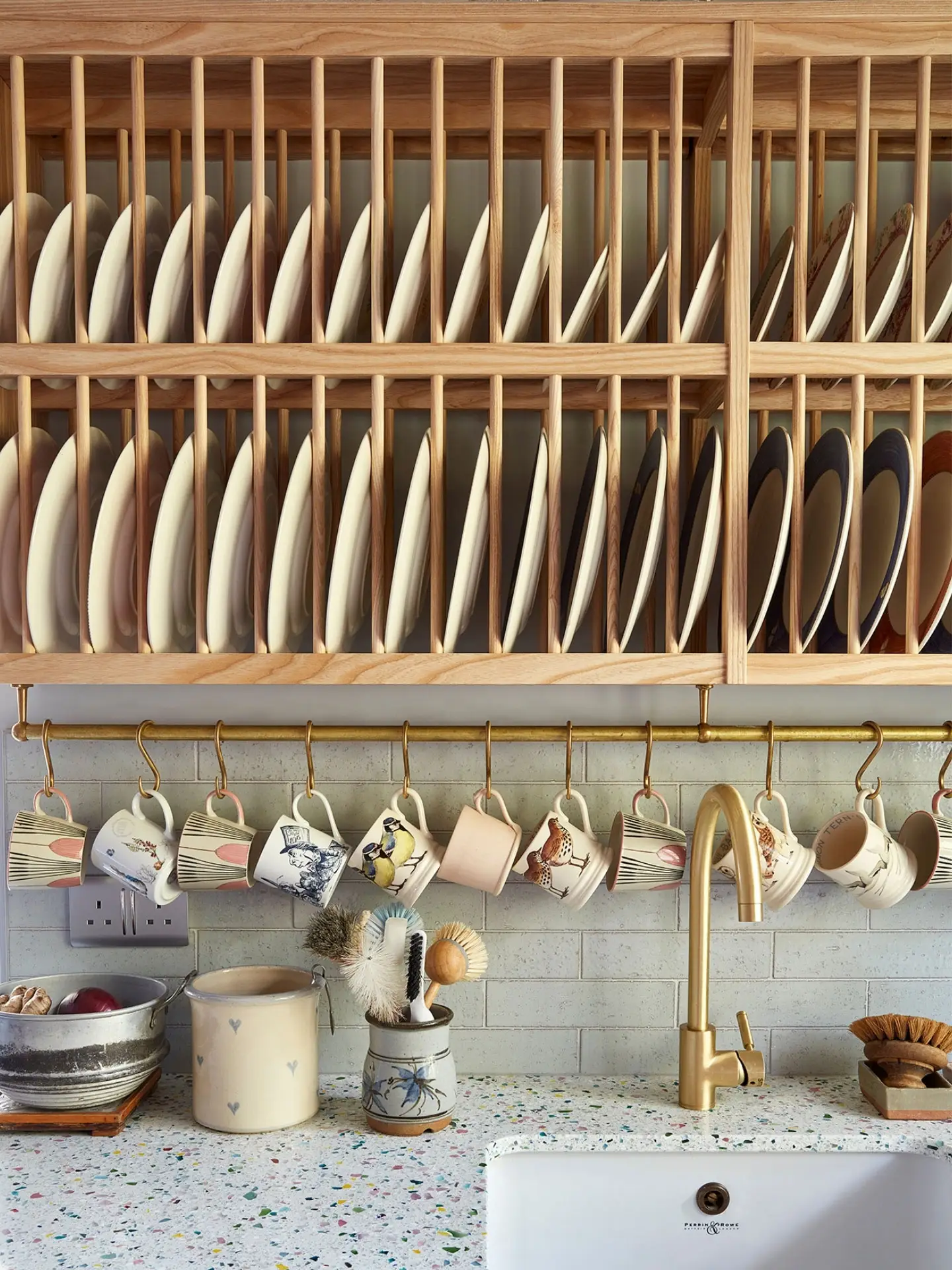 A cosy kitchen with colourful plates neatly arranged in a wooden plate rack, hanging mugs, terrazzo countertop, floral wallpaper, and a farmhouse sink.