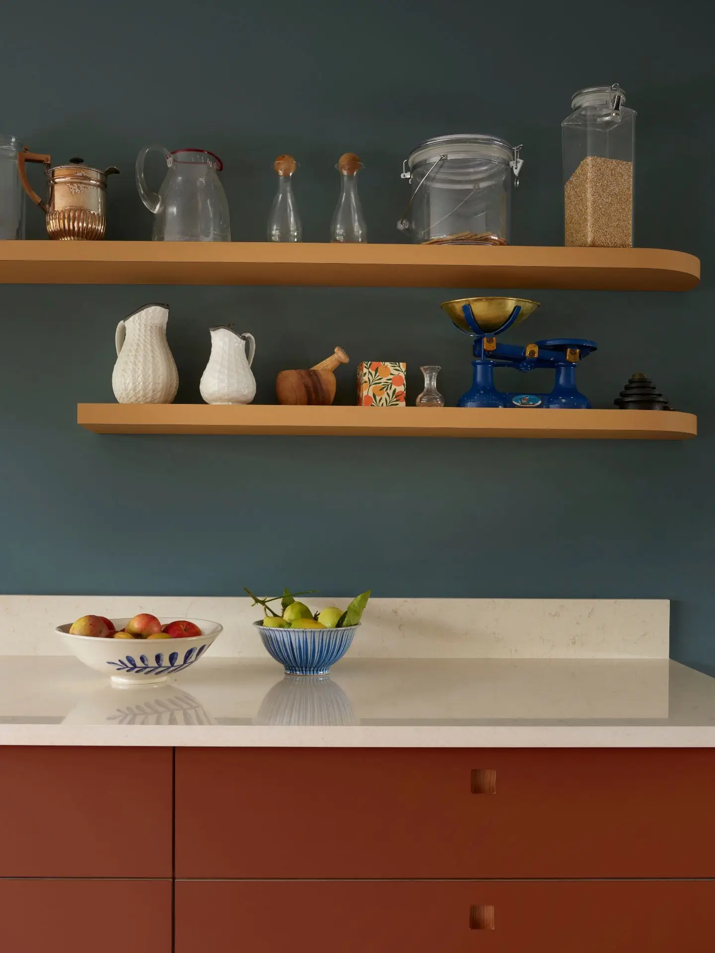 A minimalist kitchen with dark blue walls features two floating shelves. Items on display include pitchers, jars, bottles, and a blue scale. The white countertop below holds bowls of apples and pears.